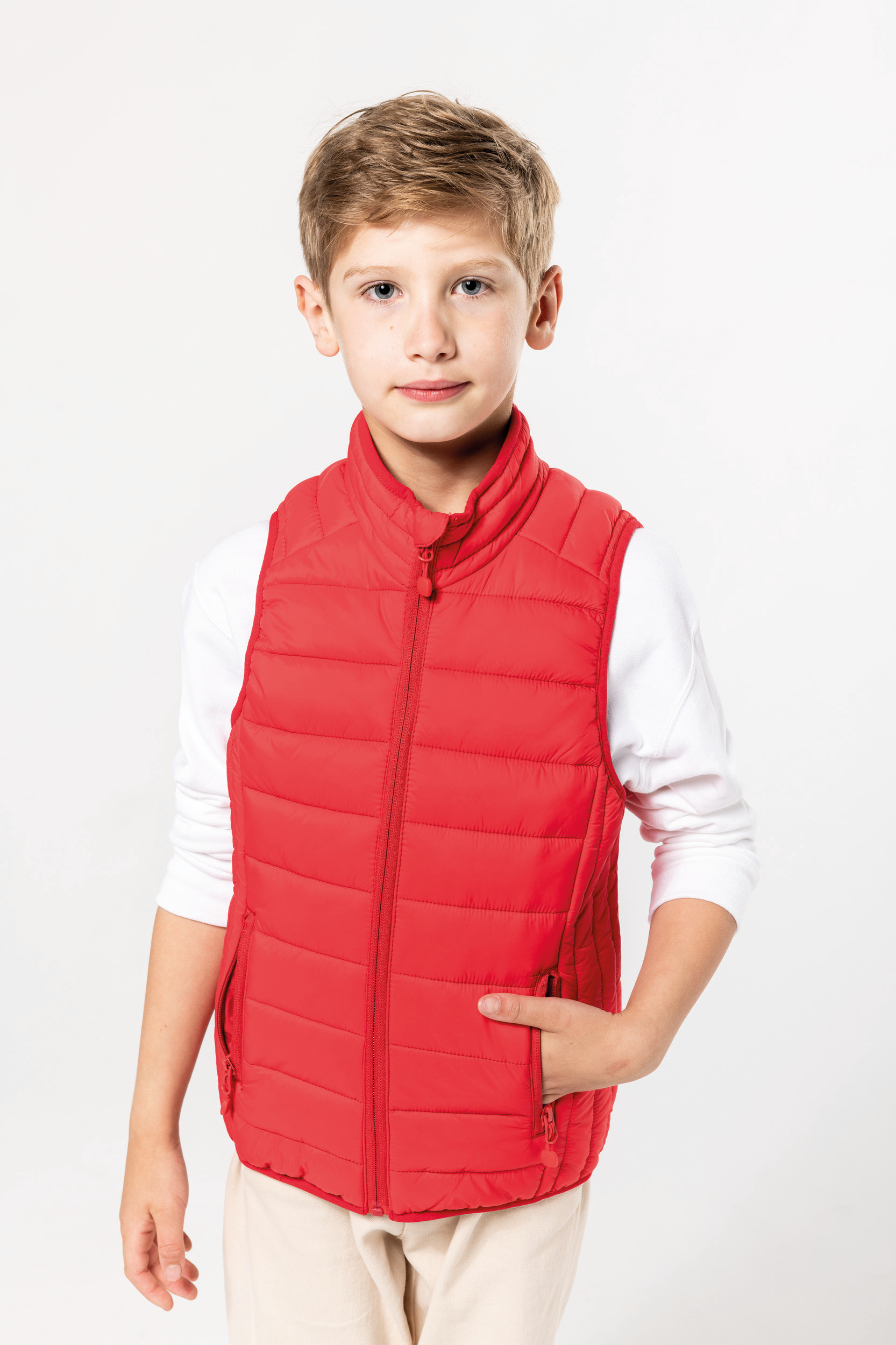 niño de pie con camiseta blanca y chaleco acolchado rojo sobre fondo neutro