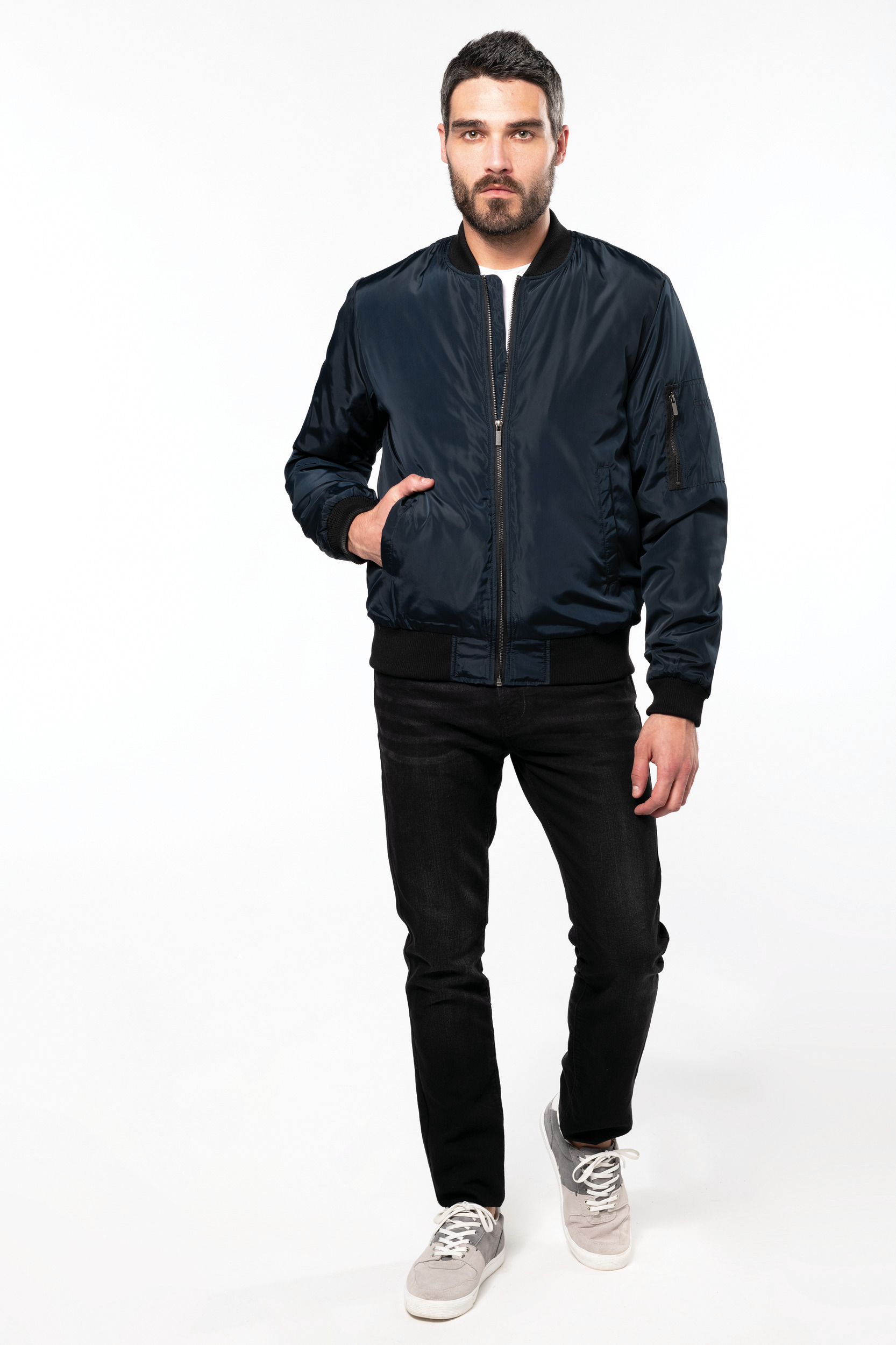 Hombre con chaqueta bomber azul marino, vaqueros negros y zapatillas grises sobre fondo blanco.