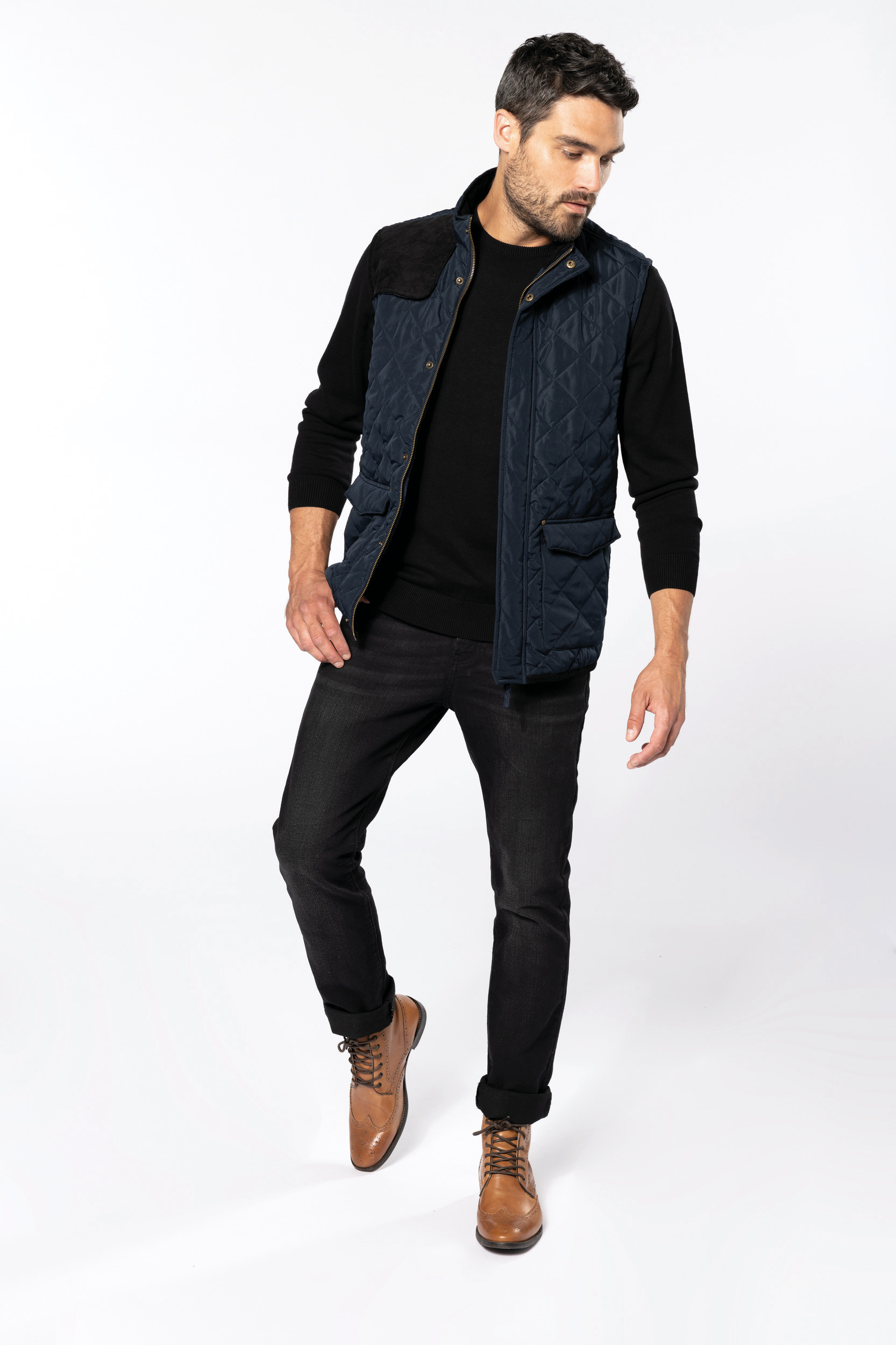 hombre con chaleco acolchado azul marino, suéter negro, vaqueros oscuros y botas marrones sobre fondo blanco