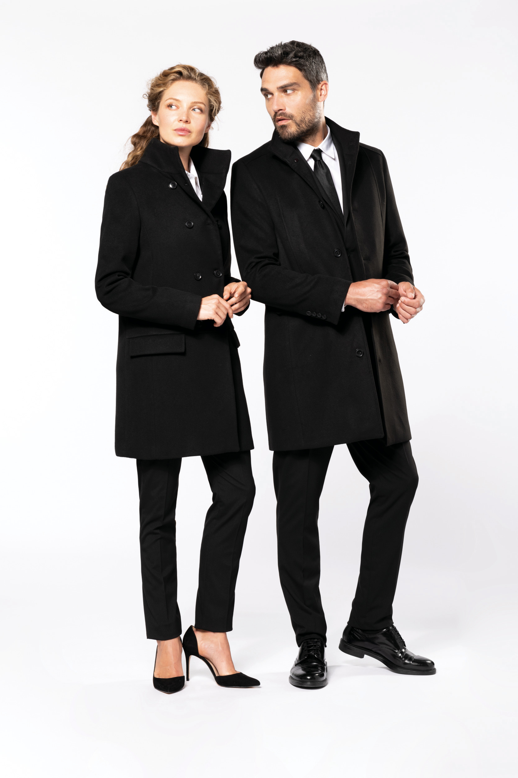 Pareja con abrigos negros de corte entallado sobre fondo blanco en postura elegante.