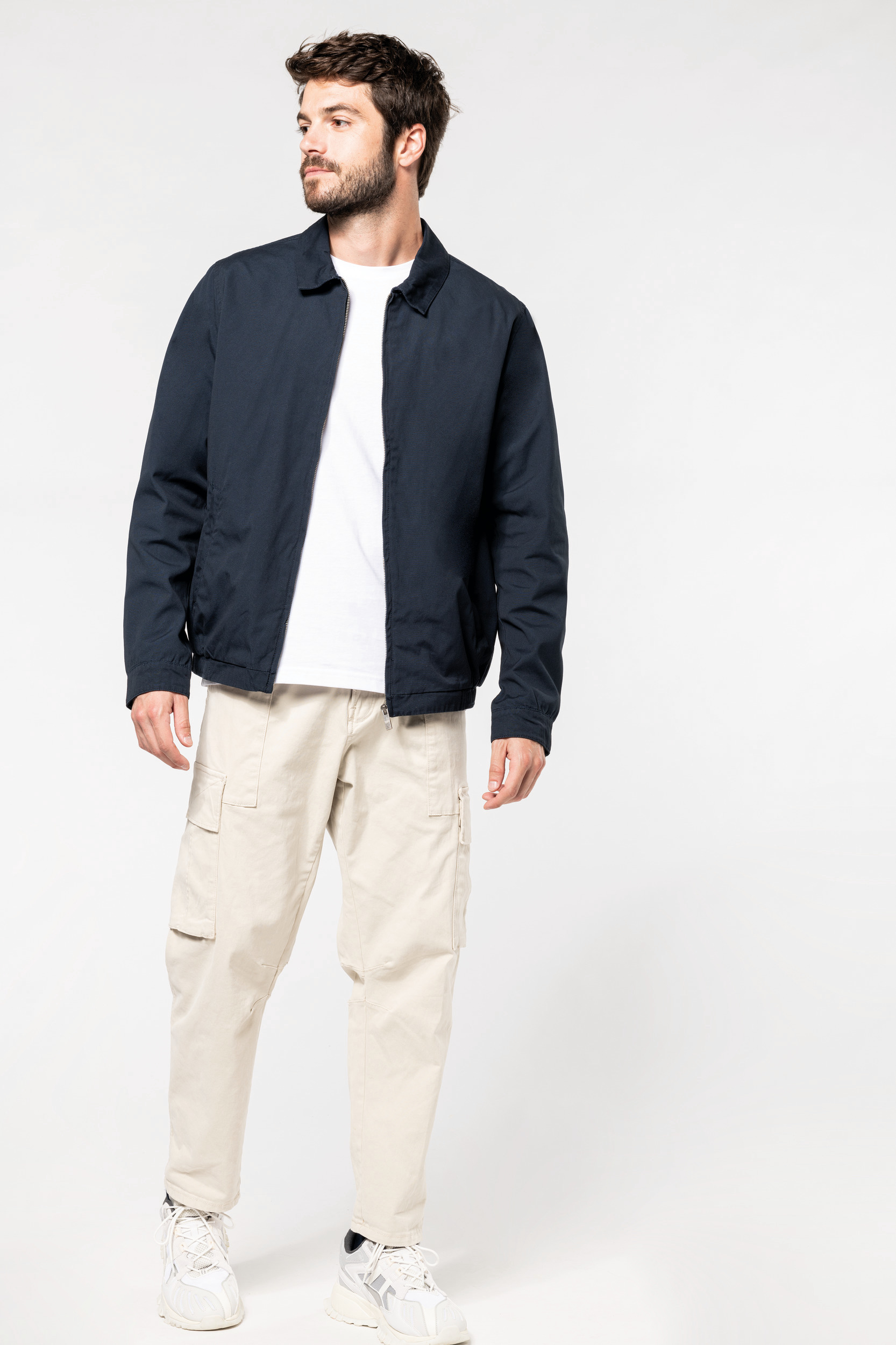 Hombre joven con cazadora azul marino, camiseta blanca y pantalones cargo beige sobre fondo blanco.