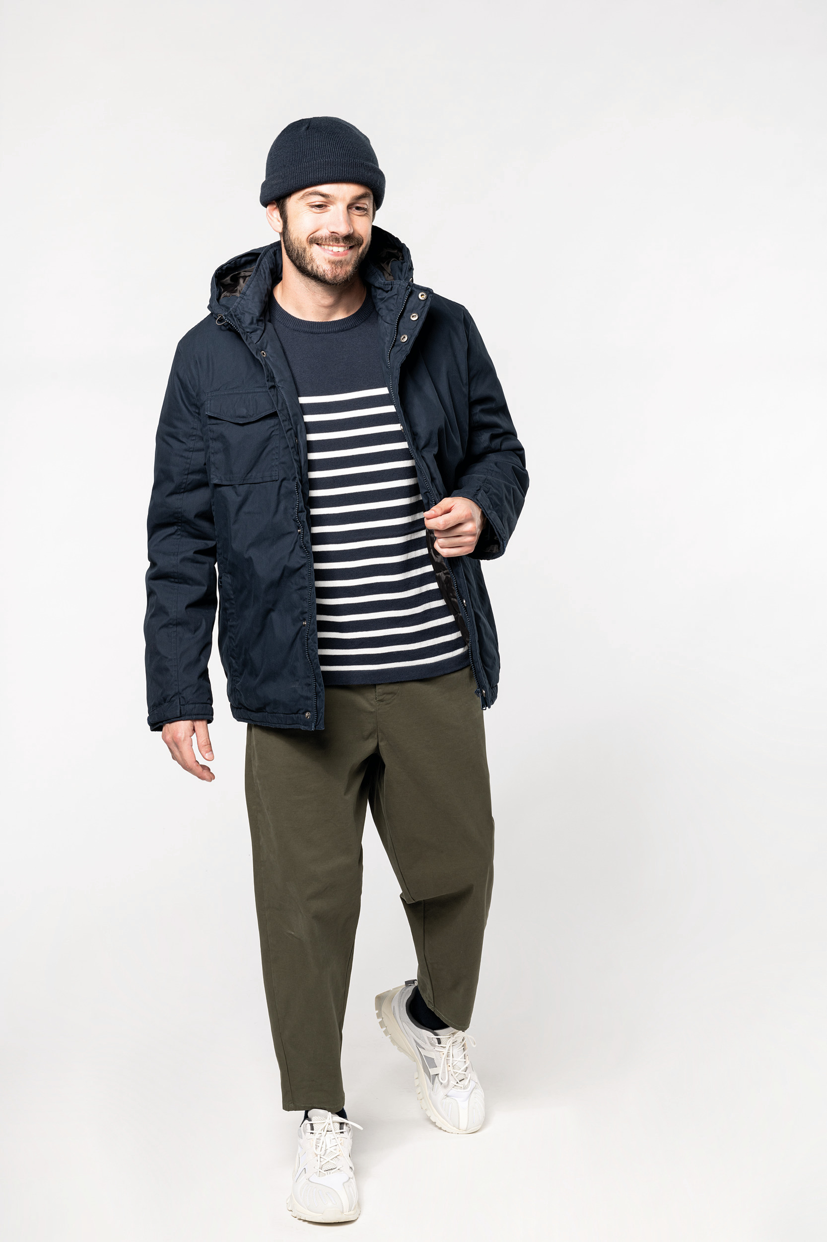 Hombre sonriente con chaqueta azul marino, jersey de rayas blancas y azul, pantalones verdes y zapatillas blancas sobre fondo blanco.