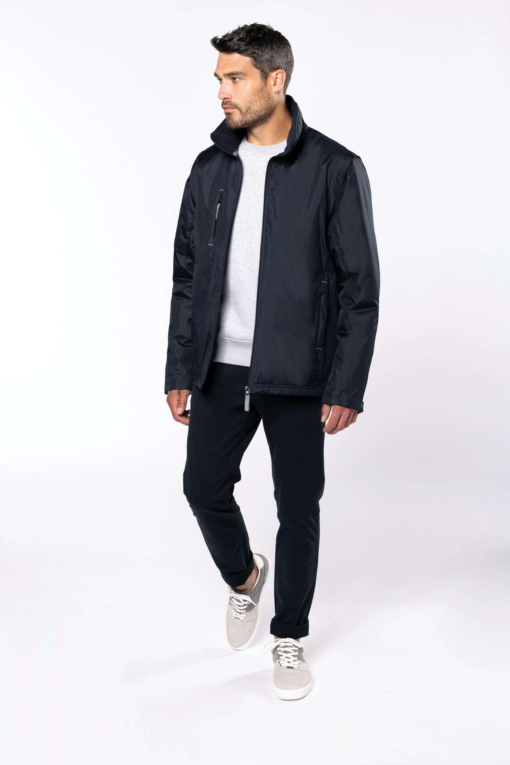 Hombre caminando con chaqueta azul marino, sudadera gris y pantalones oscuros sobre fondo blanco