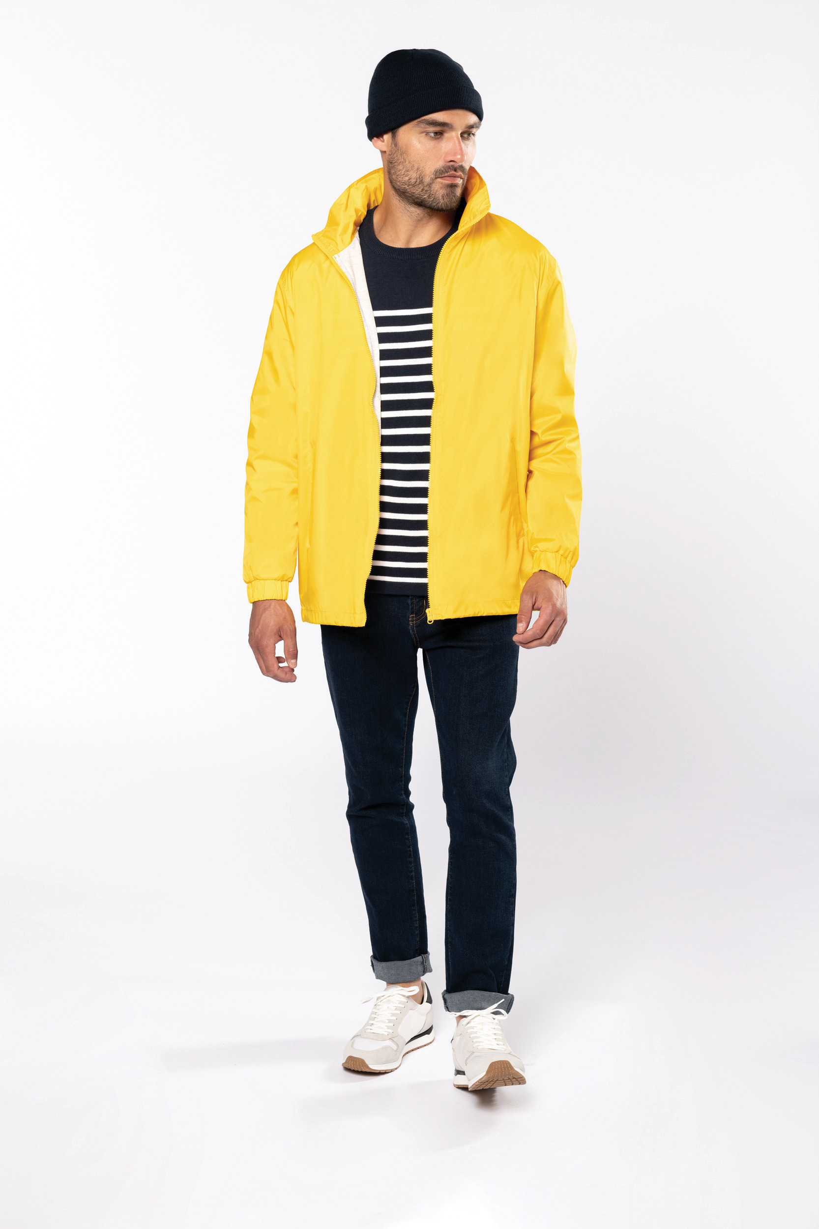 Hombre con cazadora amarilla, jersey de rayas, vaqueros oscuros y gorro beanie sobre fondo blanco.