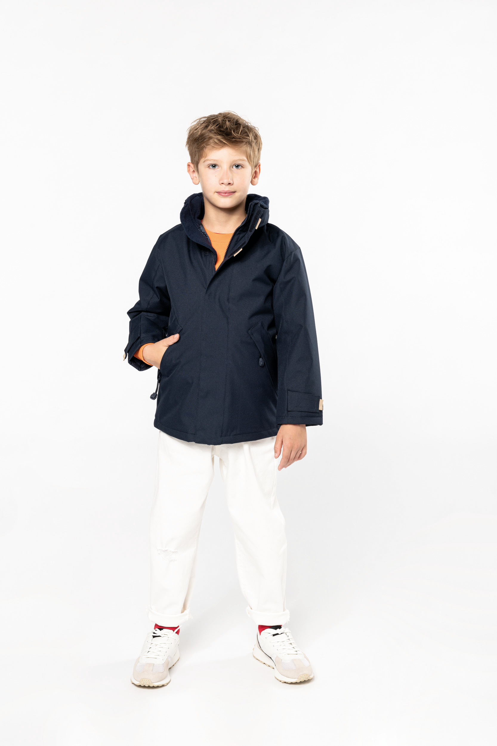 Niño de pie en estudio vistiendo parka azul marino, camiseta naranja y pantalones blancos.