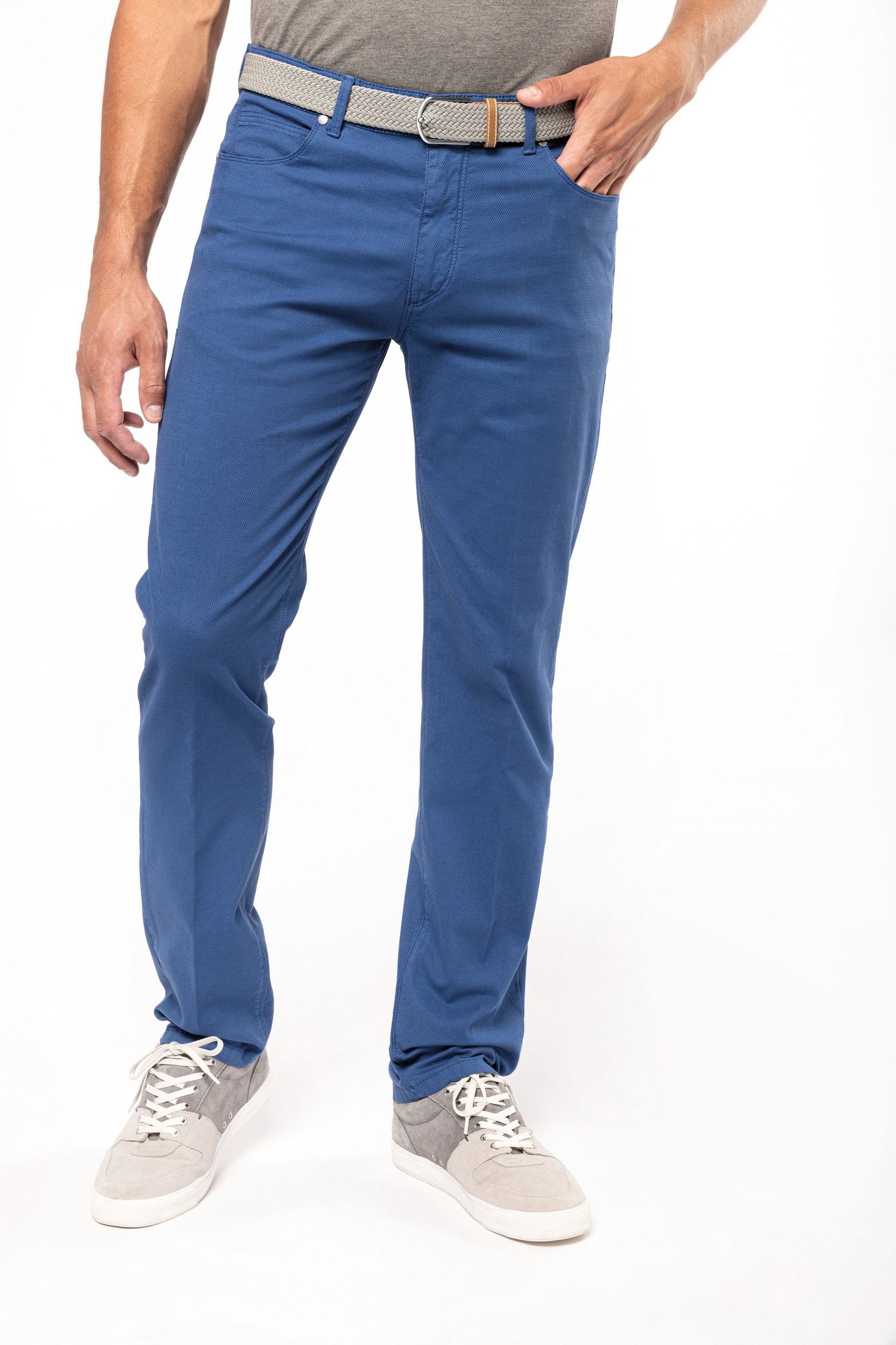 Modelo masculino con pantalones chinos azules, cinturón trenzado gris y zapatillas deportivas claras sobre fondo blanco.