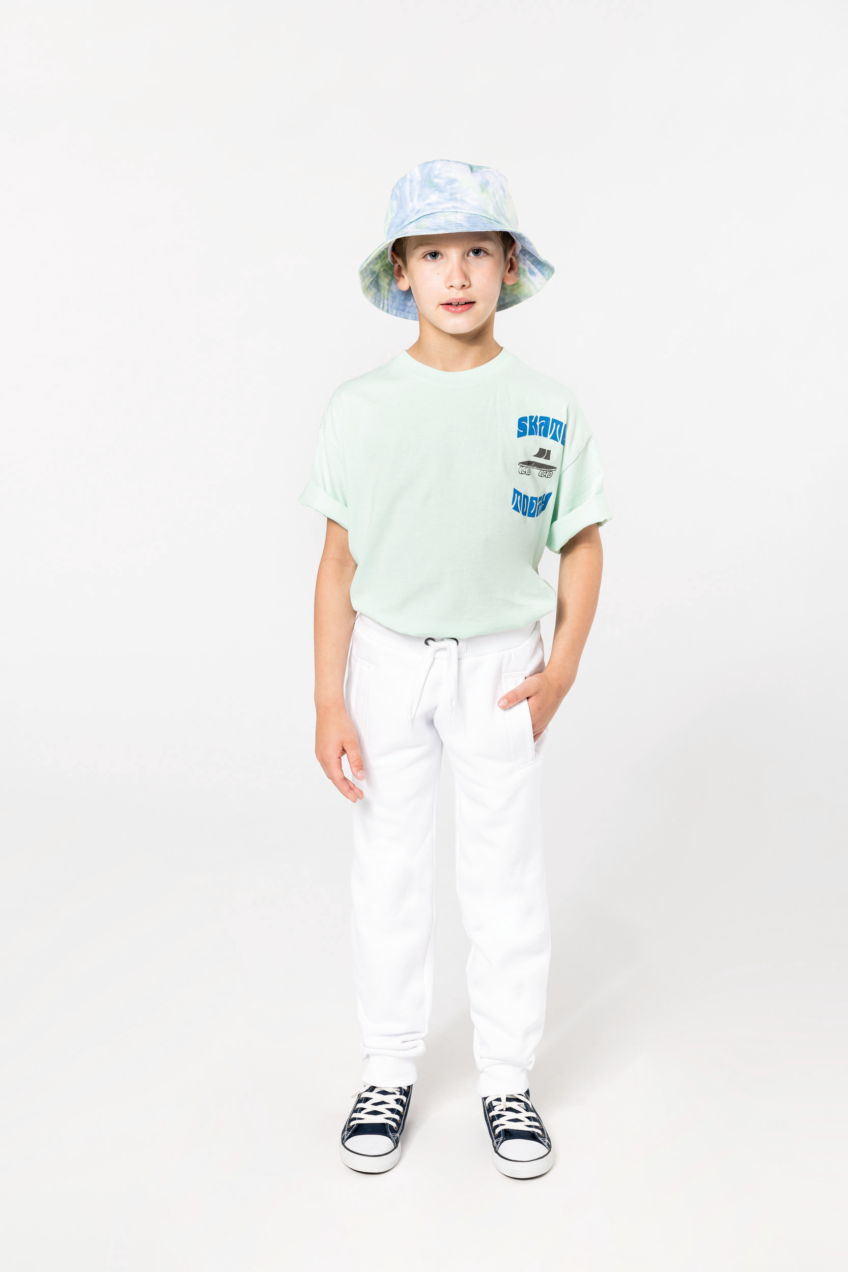 Niño de pie con camiseta verde mint, pantalones blancos, zapatillas negras y sombrero de pescador sobre fondo blanco