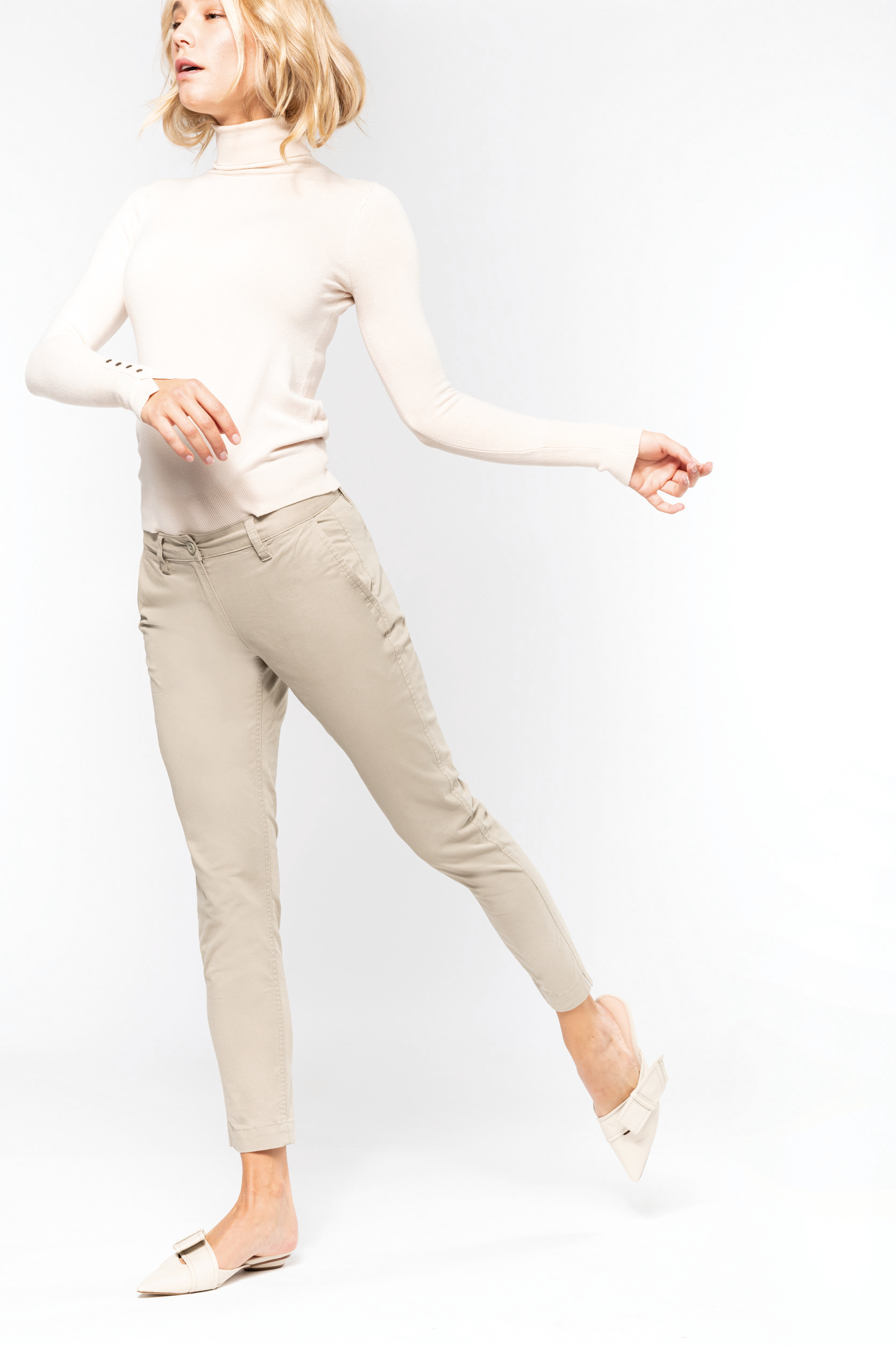 Mujer joven en estudio con pantalones chinos beige, jersey de cuello alto crema y mocasines ligeros, posando en movimiento sobre fondo blanco.