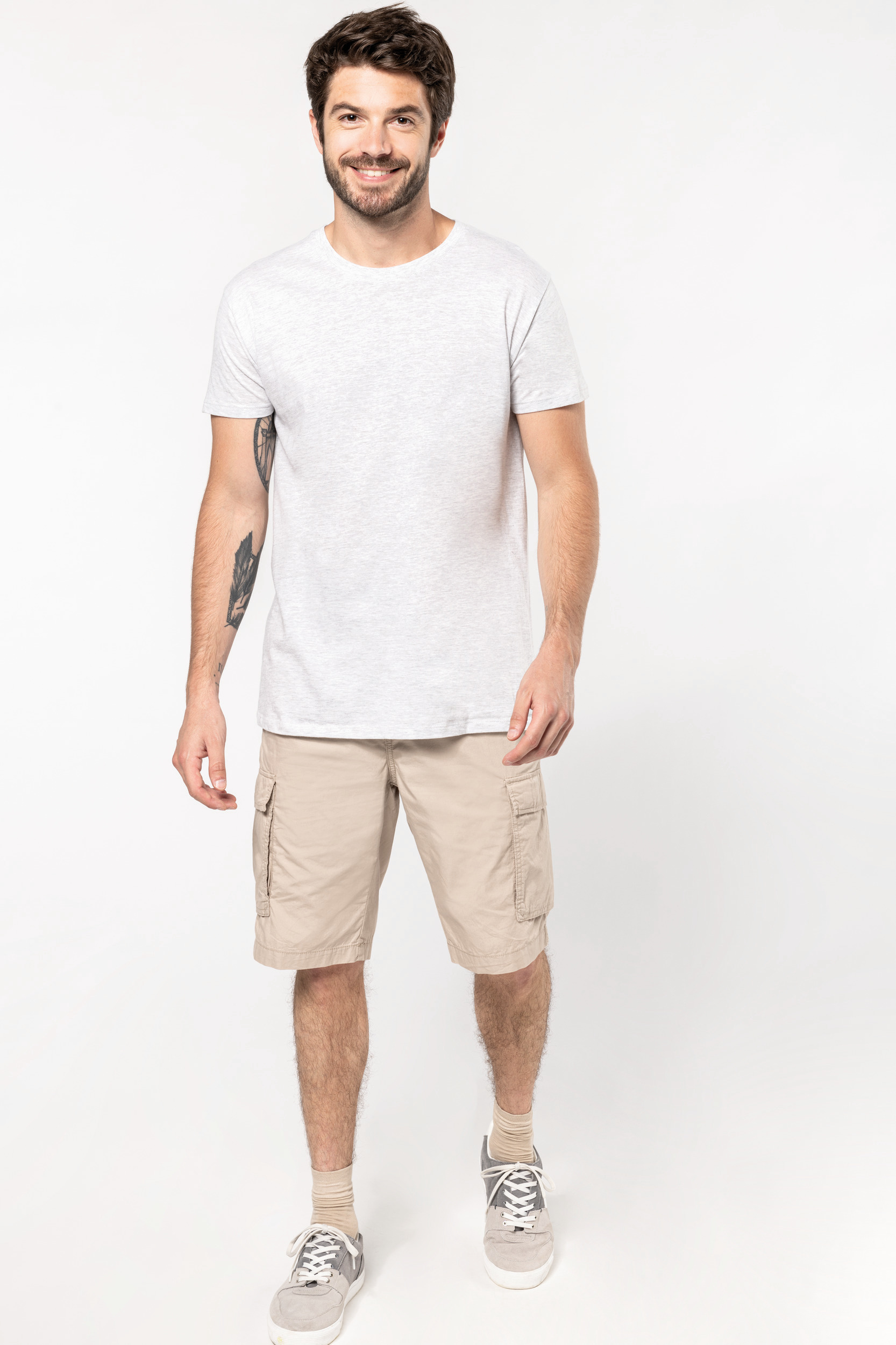 Hombre sonriente de pie con camiseta blanca, pantalones cortos cargo beige y zapatillas deportivas sobre fondo blanco.