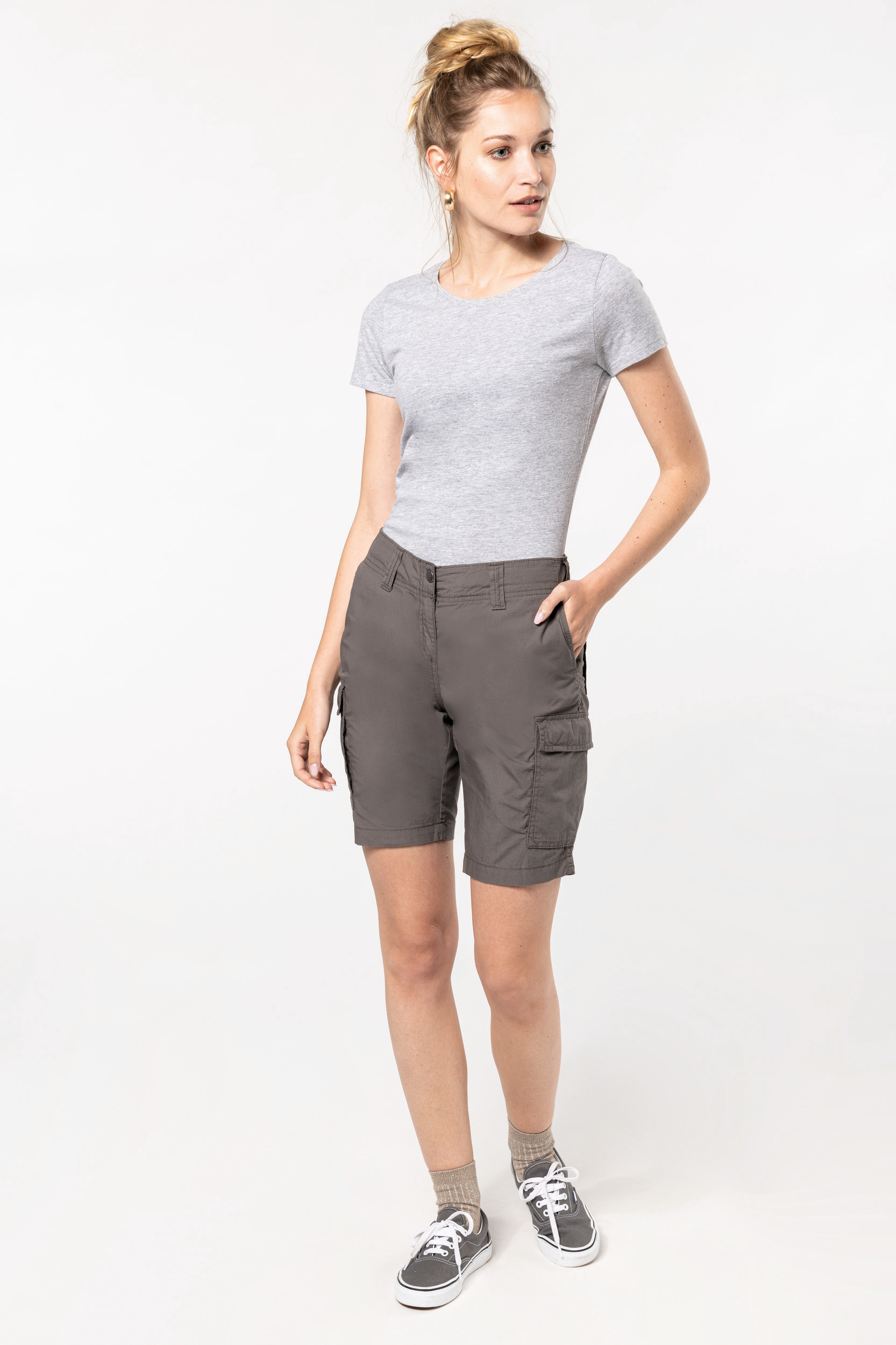 Mujer joven con camiseta gris y bermudas cargo posando sobre fondo blanco