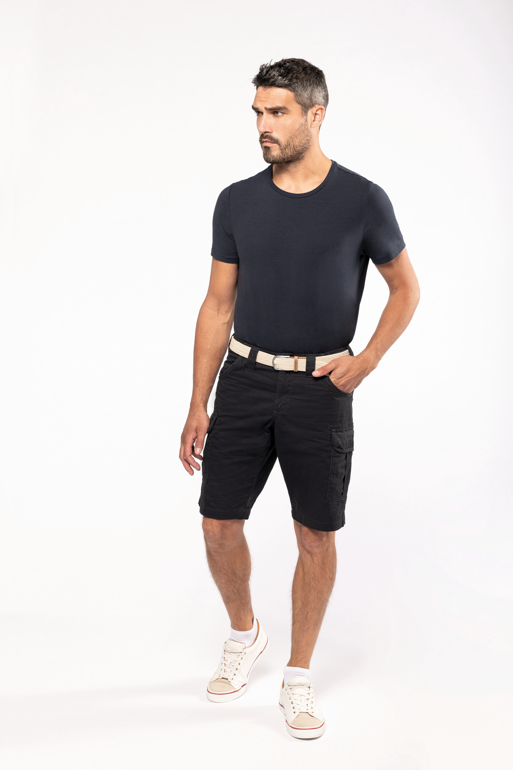 Hombre posando con camiseta negra, bermudas cargo y zapatillas blancas sobre fondo blanco