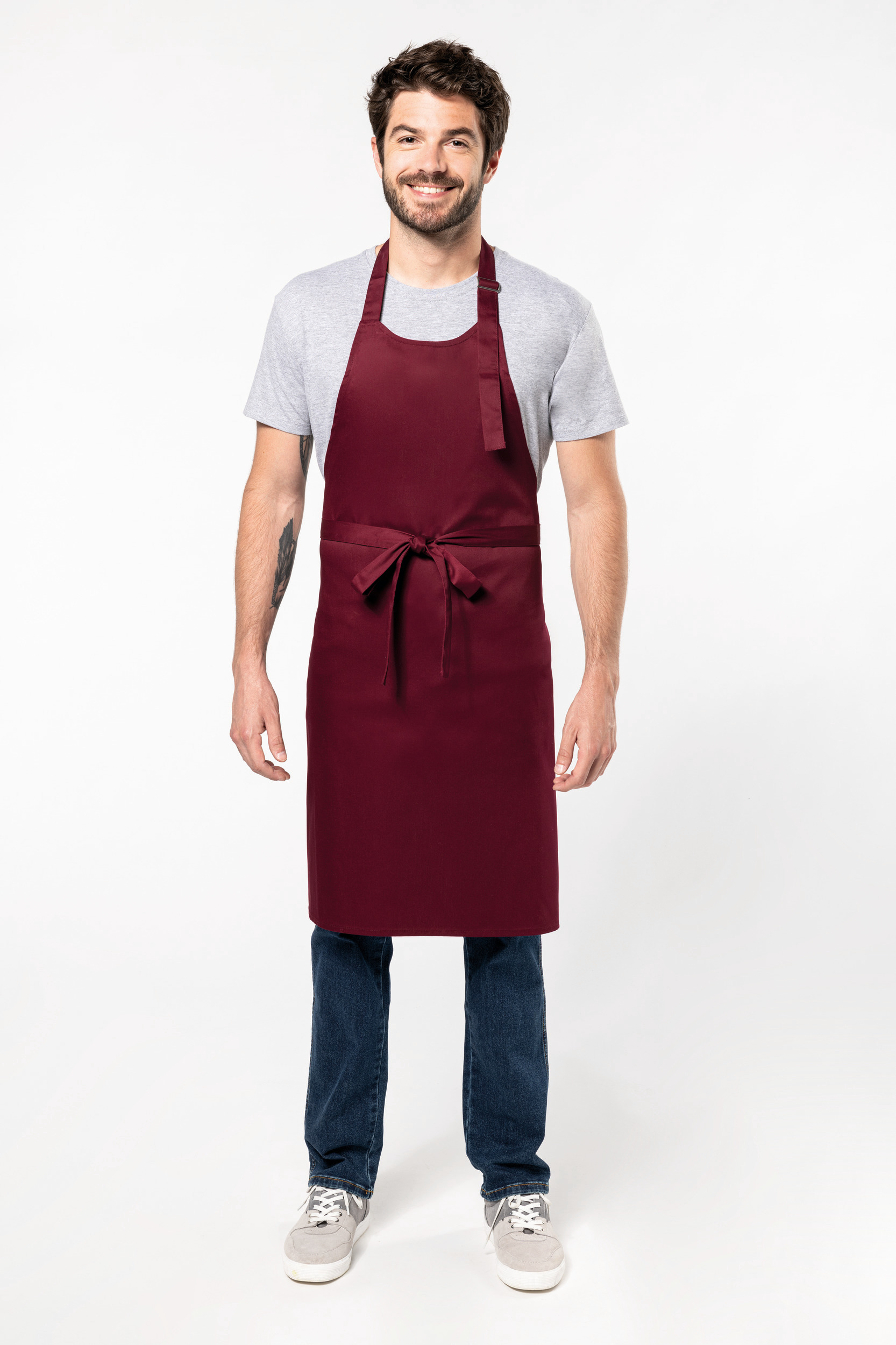 Hombre sonriente con delantal granate sobre camiseta gris y vaqueros, fondo blanco.