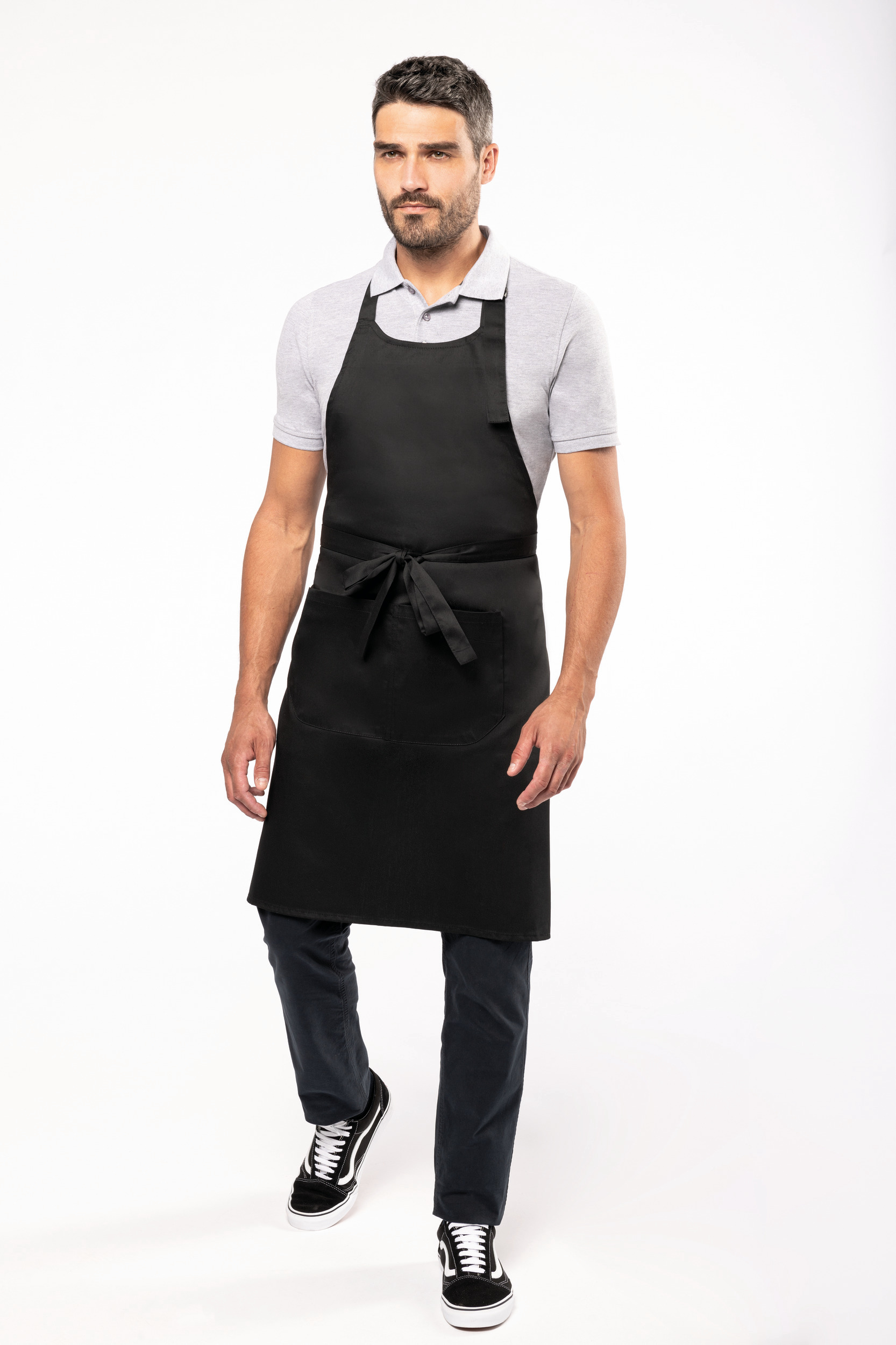 Hombre de pie con polo gris y delantal negro atado en la cintura sobre fondo blanco.