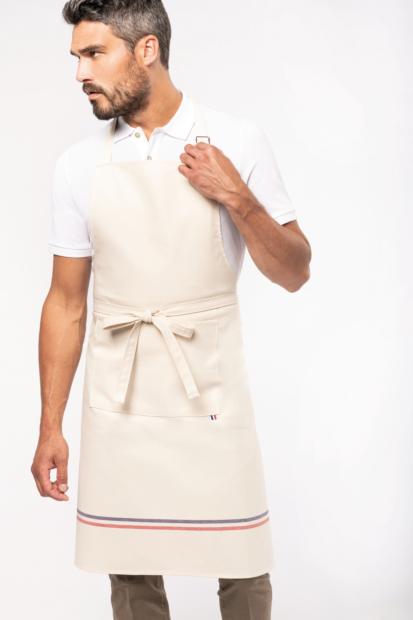 Hombre con delantal crema de algodón y bolsillo frontal, ajustando la cinta del cuello sobre camisa polo blanca.