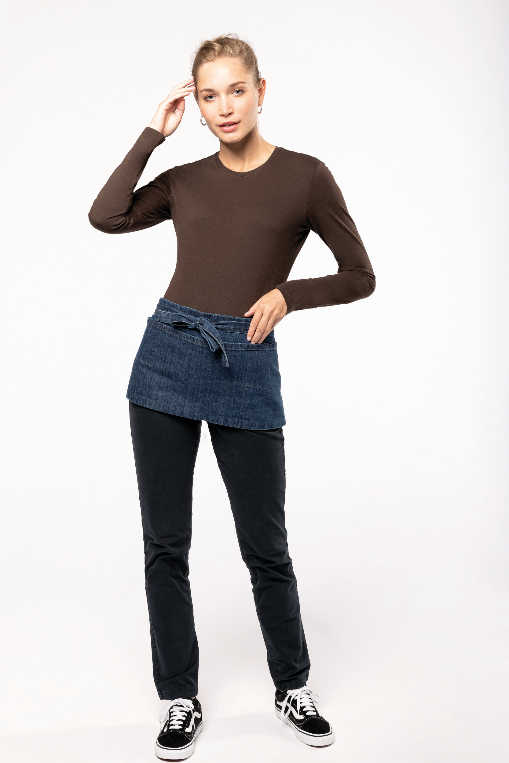 Mujer joven con camiseta de manga larga marrón y delantal vaquero anudado sobre pantalones negros y zapatillas deportivas negras y blancas