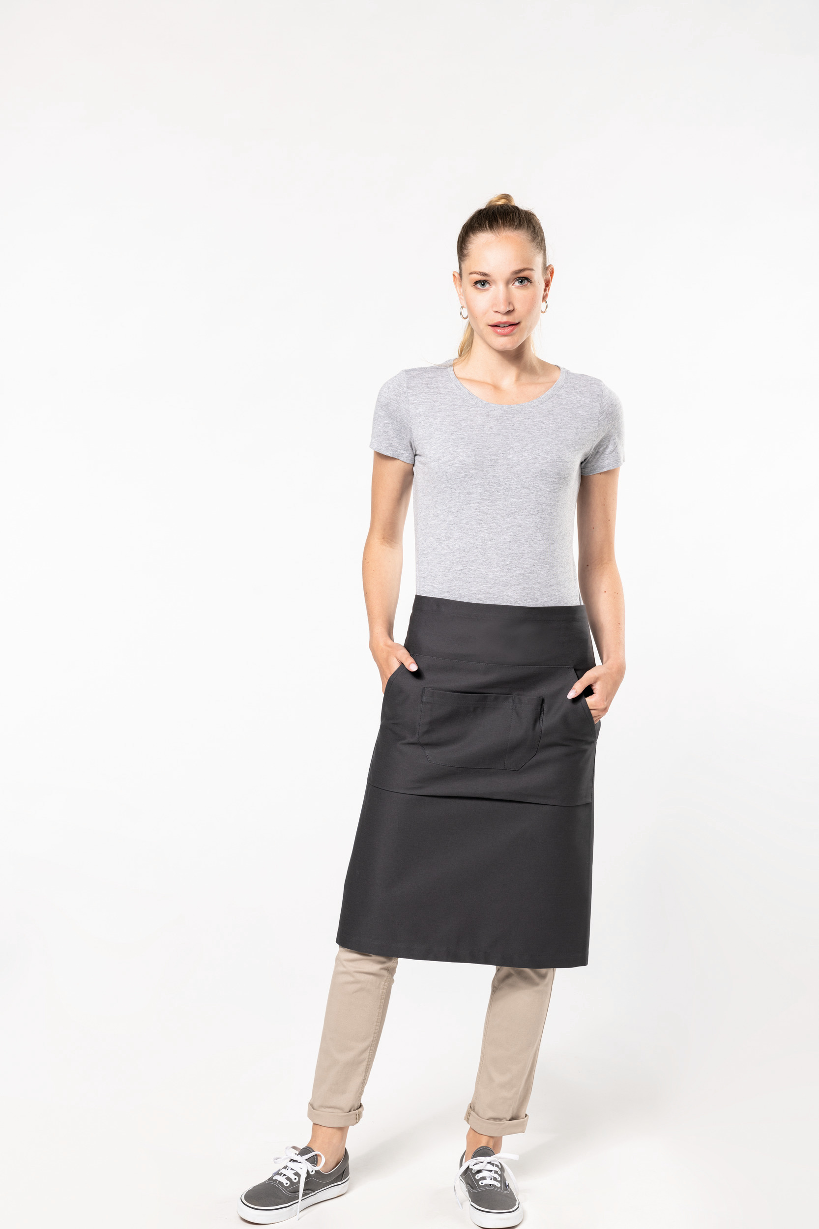 Mujer joven con camiseta gris y delantal negro con bolsillos, sobre fondo blanco