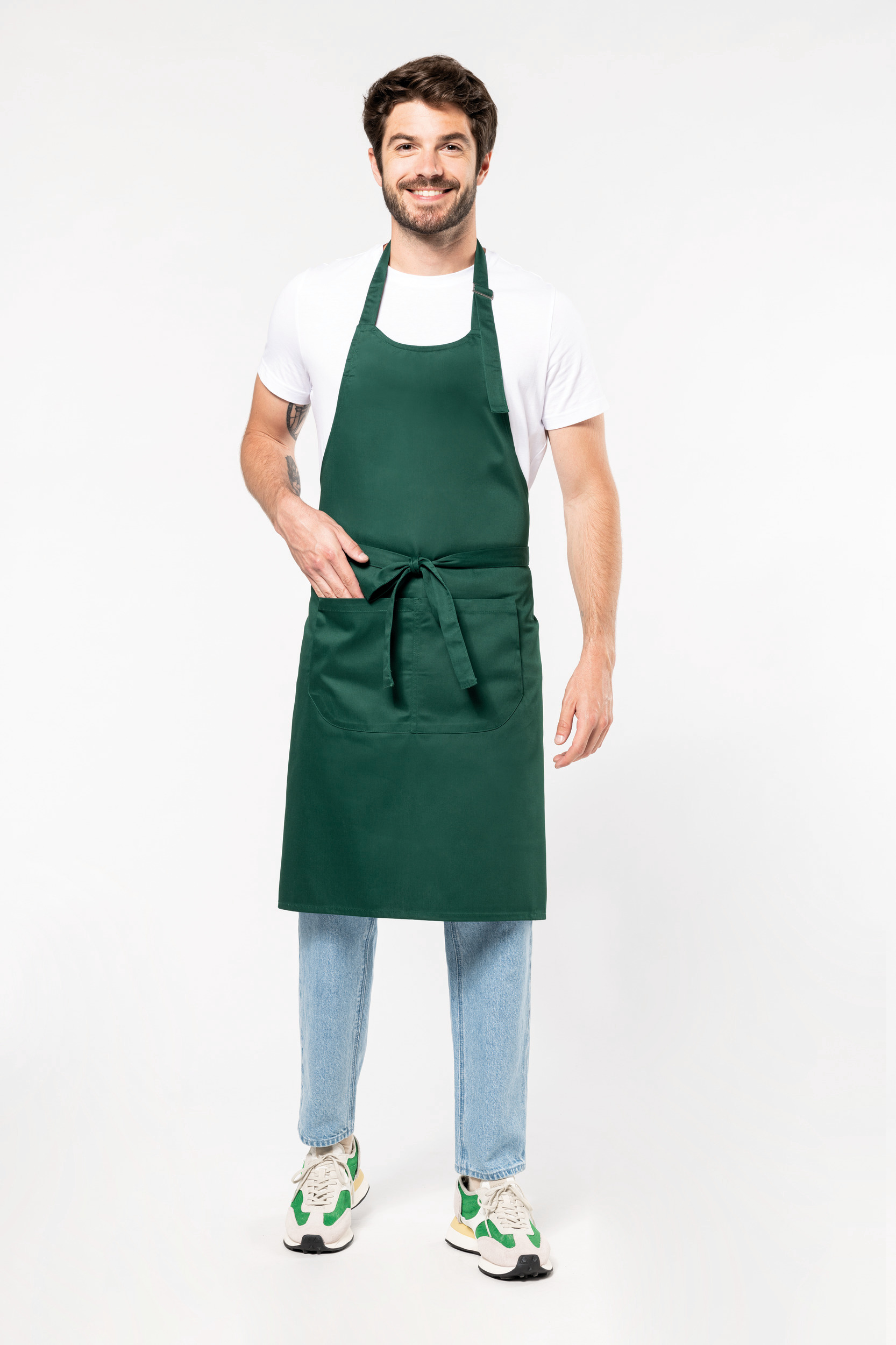 Hombre sonriente con delantal verde oscuro sobre camiseta blanca y vaqueros delante de fondo neutro.