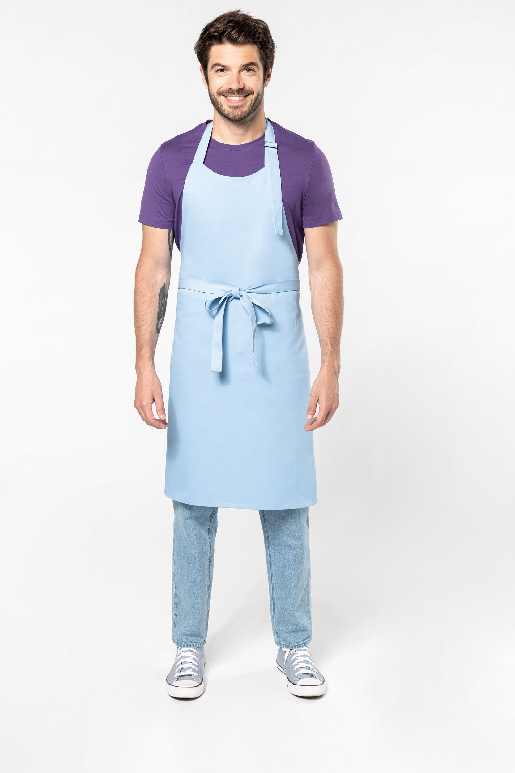 Hombre sonriente de pie con delantal azul claro sobre camiseta morada y vaqueros en fondo blanco.