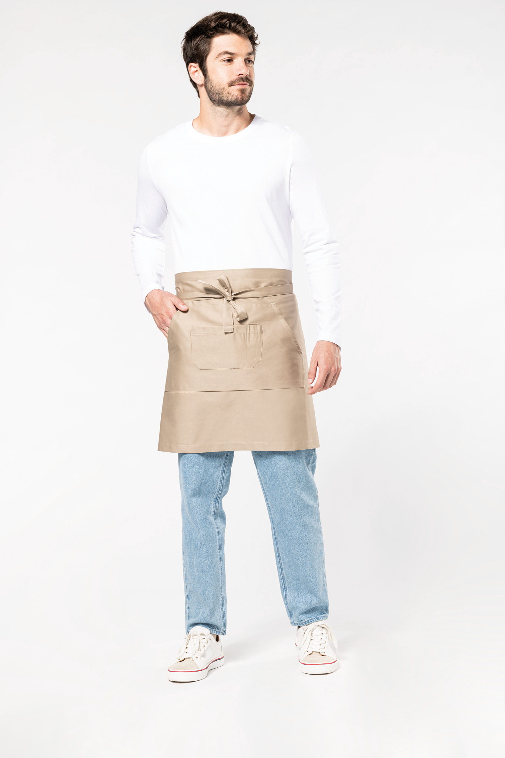 Hombre de pie con delantal beige atado a la cintura, camiseta blanca y vaqueros sobre fondo claro.