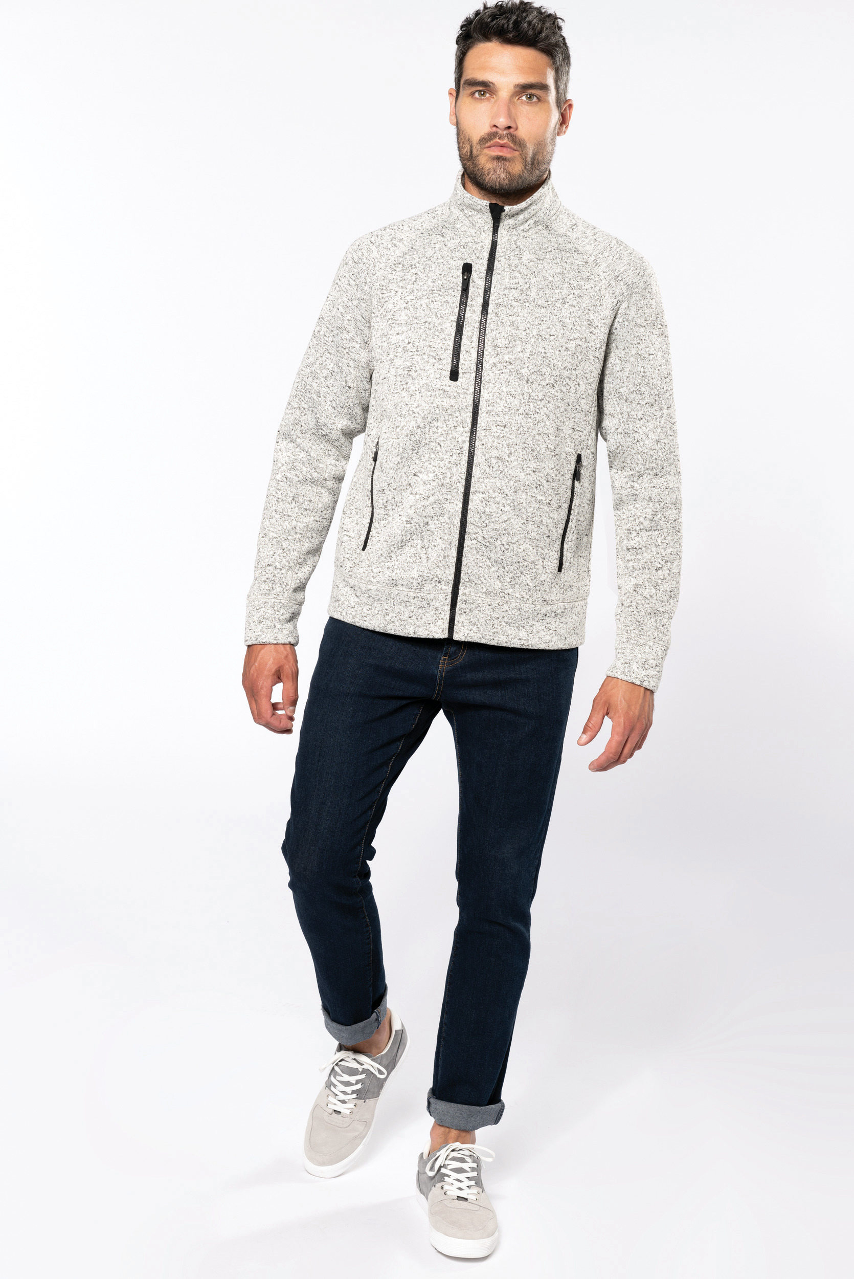 Hombre caminando con chaqueta de punto gris, vaqueros oscuros y zapatillas sobre fondo blanco