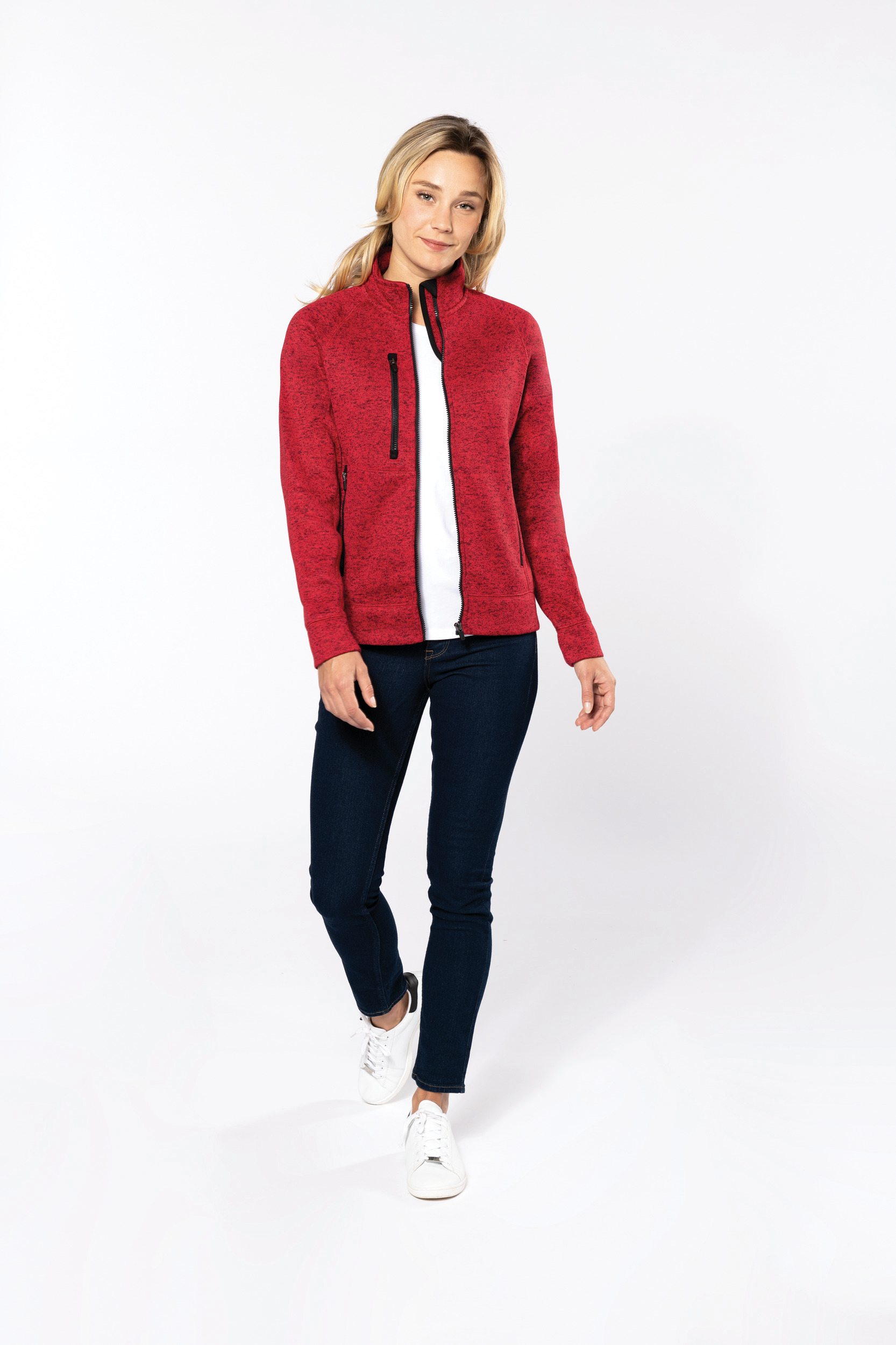 Mujer caminando con chaqueta polar roja, camiseta blanca, vaqueros ajustados y zapatillas blancas sobre fondo blanco