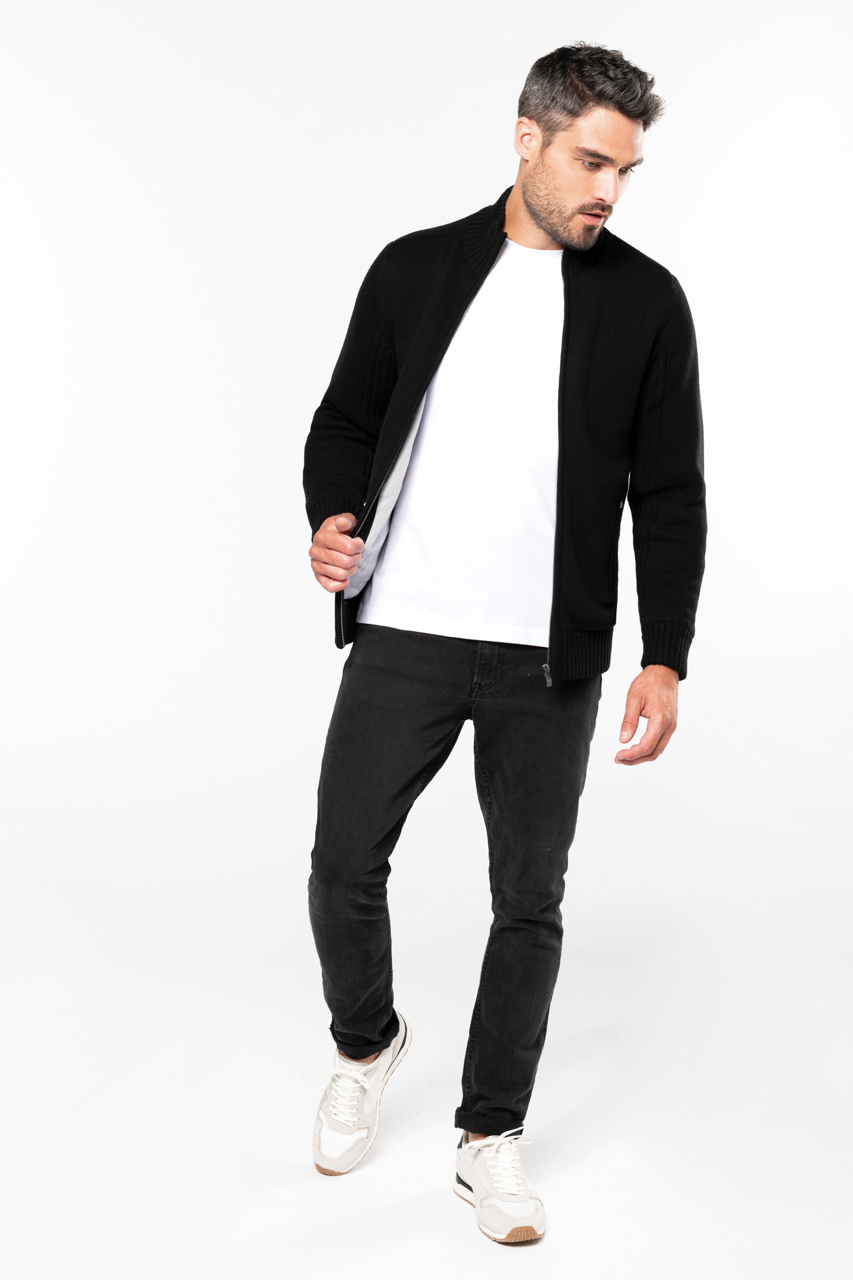 Hombre caminando con chaqueta de punto negra, camiseta blanca, vaqueros oscuros y zapatillas deportivas sobre fondo blanco