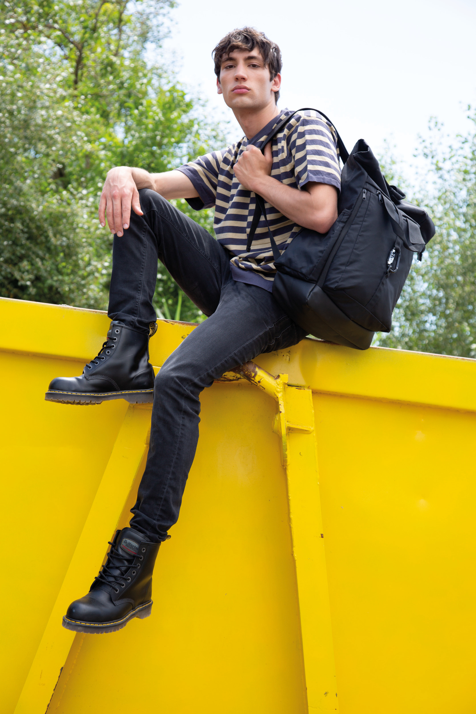 Chico con camiseta de rayas, botas negras y mochila apoyado en un contenedor amarillo al aire libre