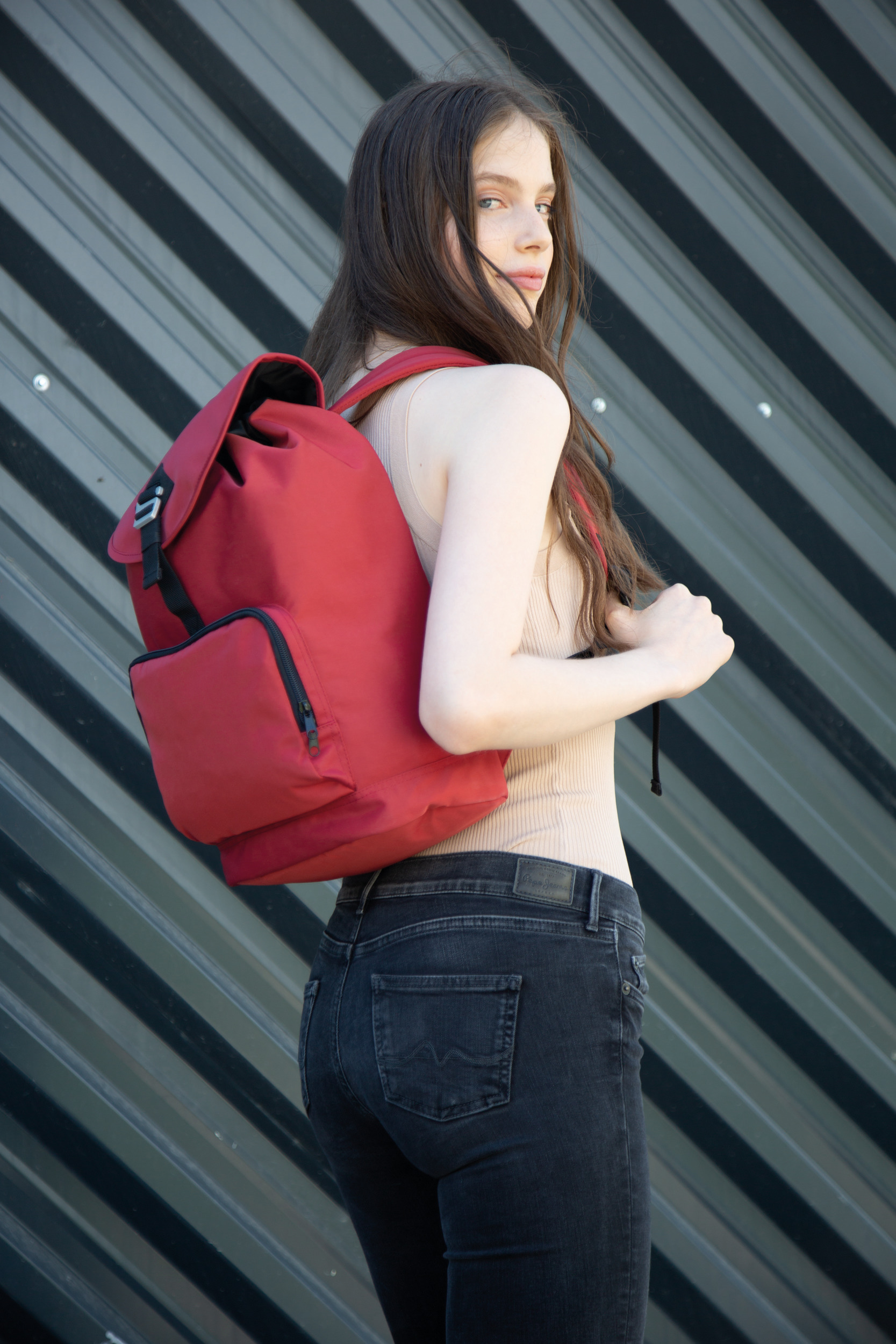 Joven de espaldas con mochila roja y top beige frente a una pared metálica de líneas diagonales.