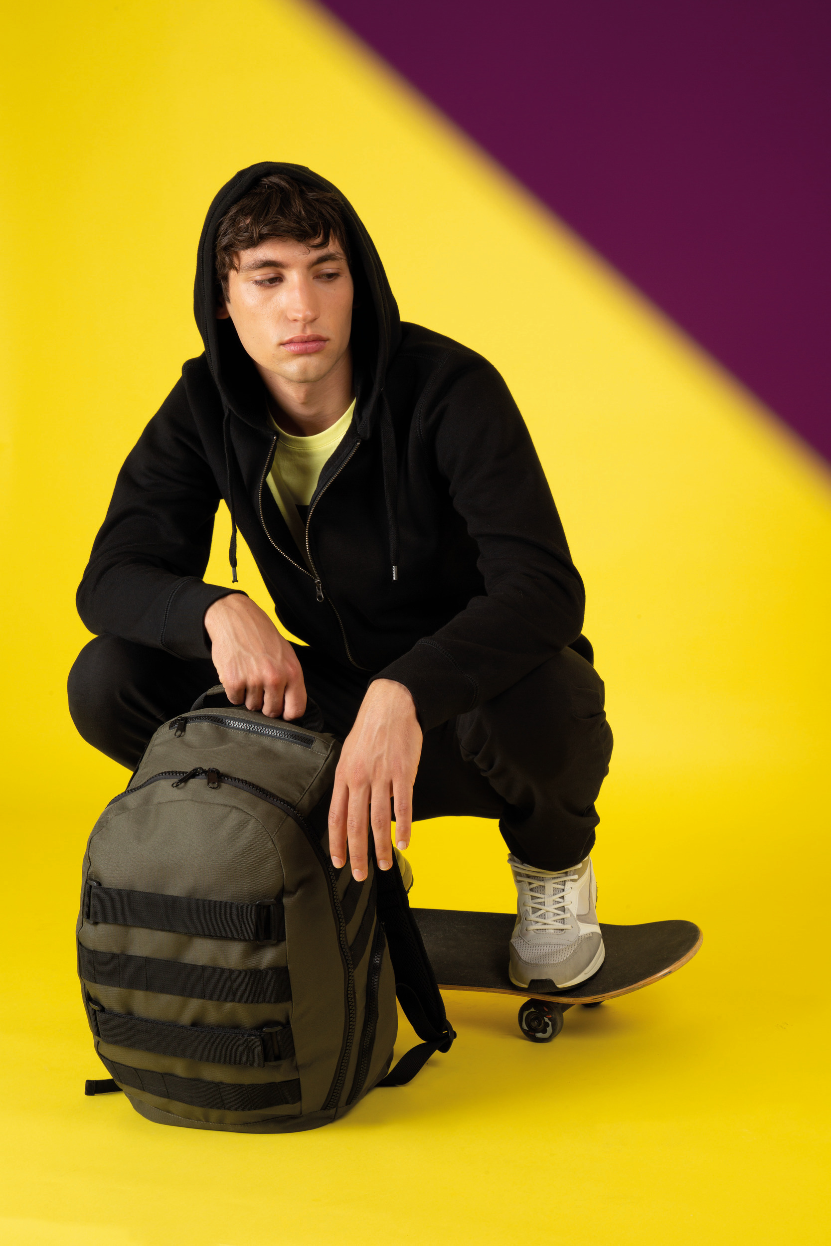 Joven con sudadera negra y zapatillas apoyado en un skate junto a una mochila sobre fondo amarillo