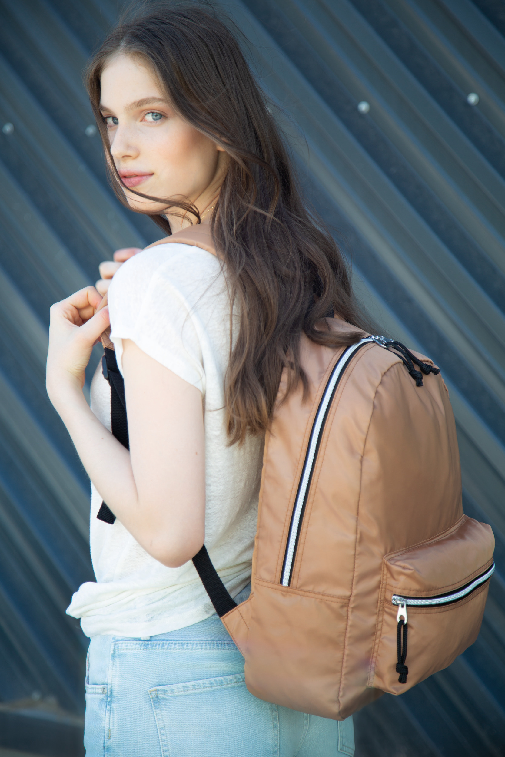 Chica joven con mochila marrón de tela, de pie junto a una pared metálica, mirando hacia la cámara.