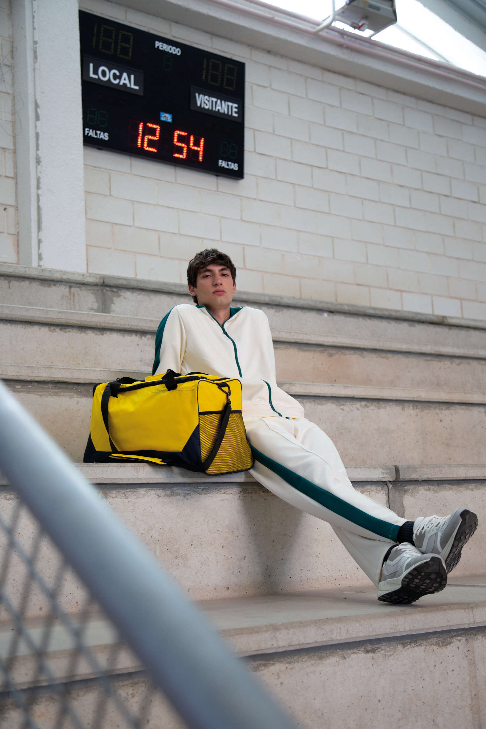 Joven con chándal blanco y verde sentado en las gradas de un polideportivo junto a una bolsa deportiva amarilla y un marcador electrónico de fondo.