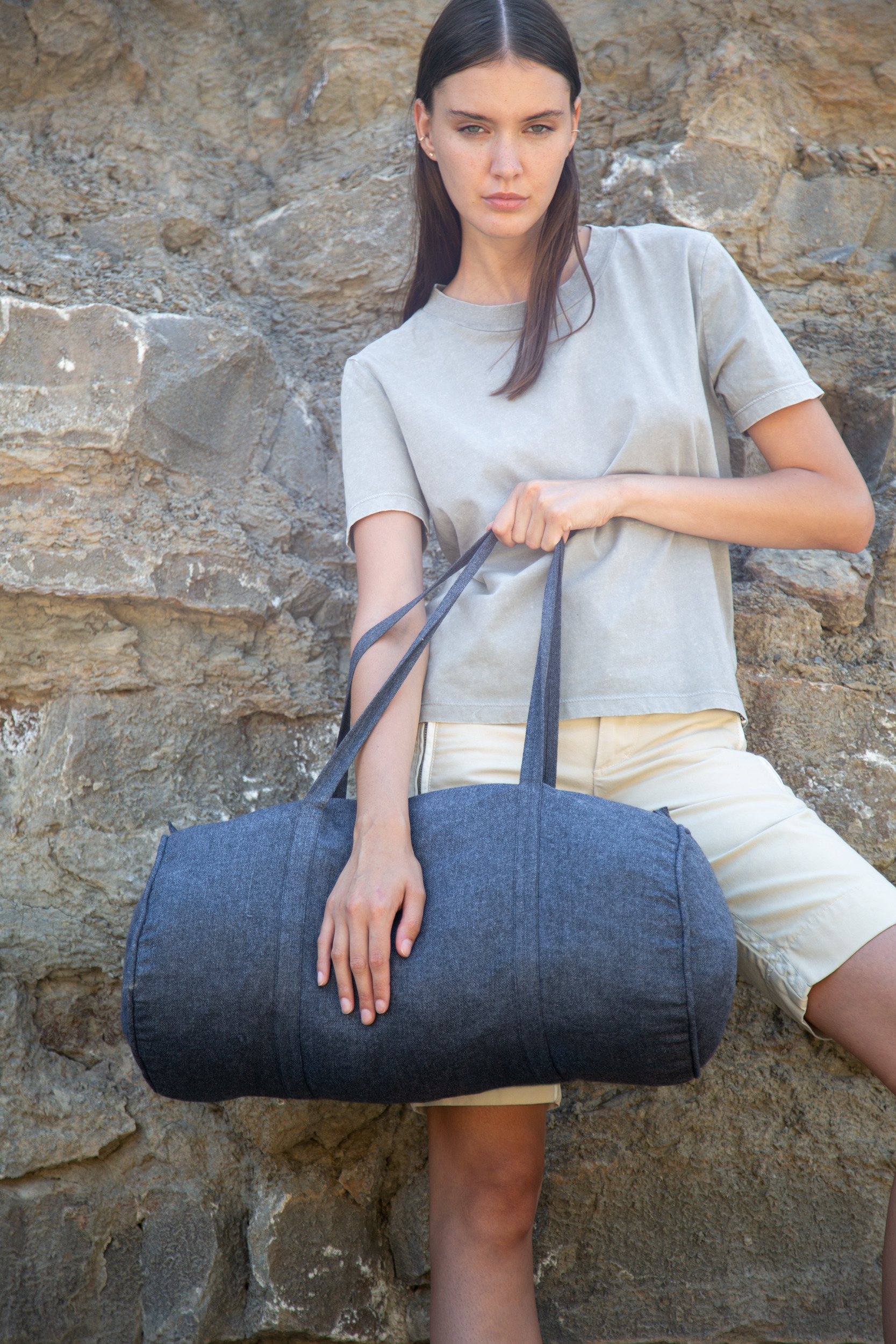 Mujer sujeta un bolso tipo saco de tela gris oscuro apoyada en un muro de piedra