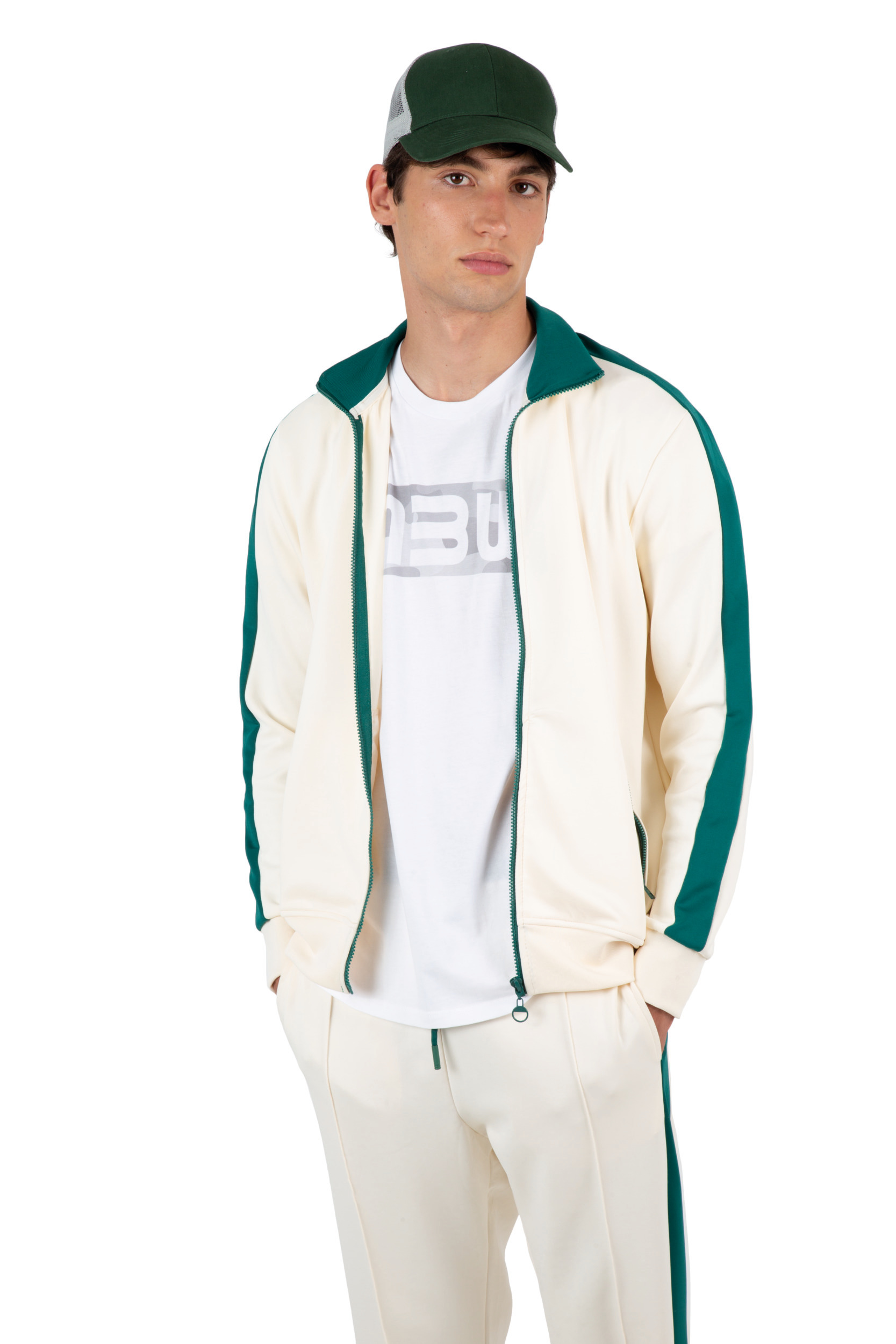 Hombre joven con chándal crema de franjas verdes, camiseta blanca y gorra verde sobre fondo blanco.