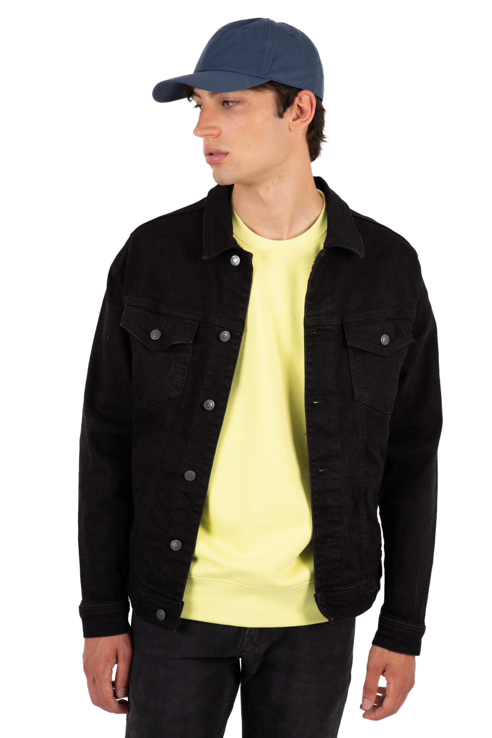 Hombre joven de perfil llevando gorra azul, sudadera amarilla y chaqueta vaquera negra.