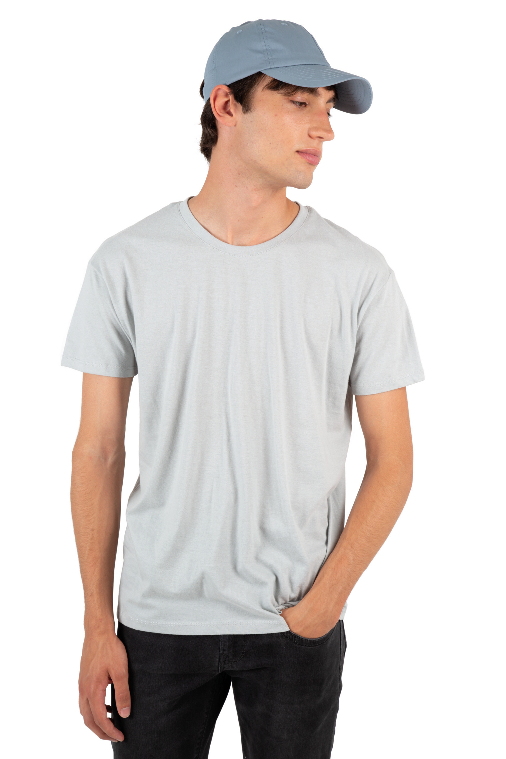 Hombre joven de pie con camiseta gris y gorra azul sobre fondo blanco, con una mano en el bolsillo.