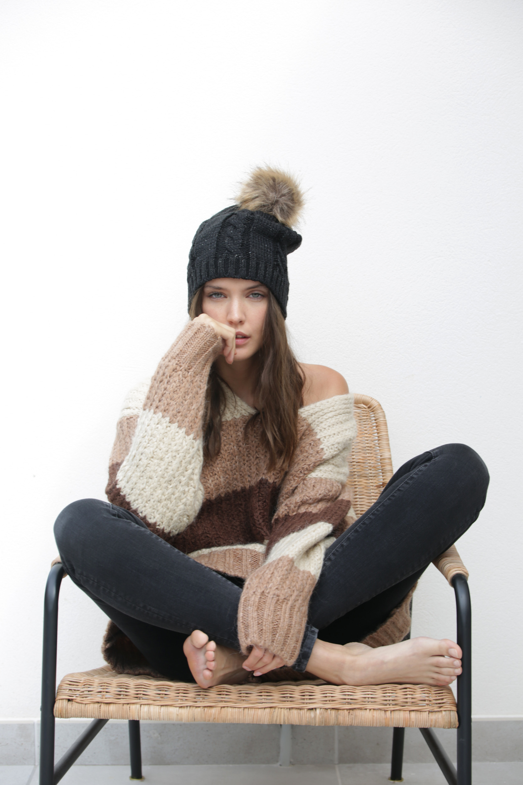 Mujer sentada de piernas cruzadas en silla de mimbre con jersey de rayas marrones y gorro de lana con pompón