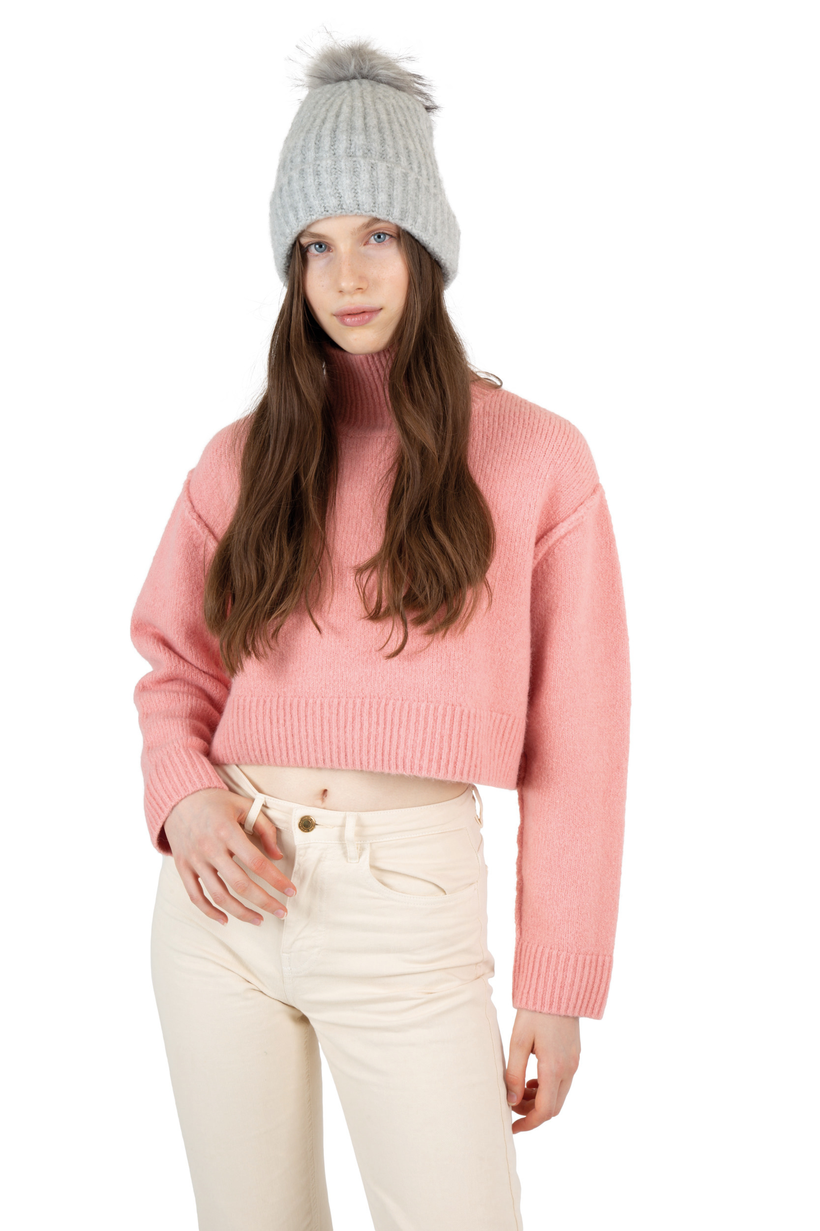Mujer joven con suéter corto rosa de punto, pantalones claros y gorro gris con pompón sobre fondo blanco.