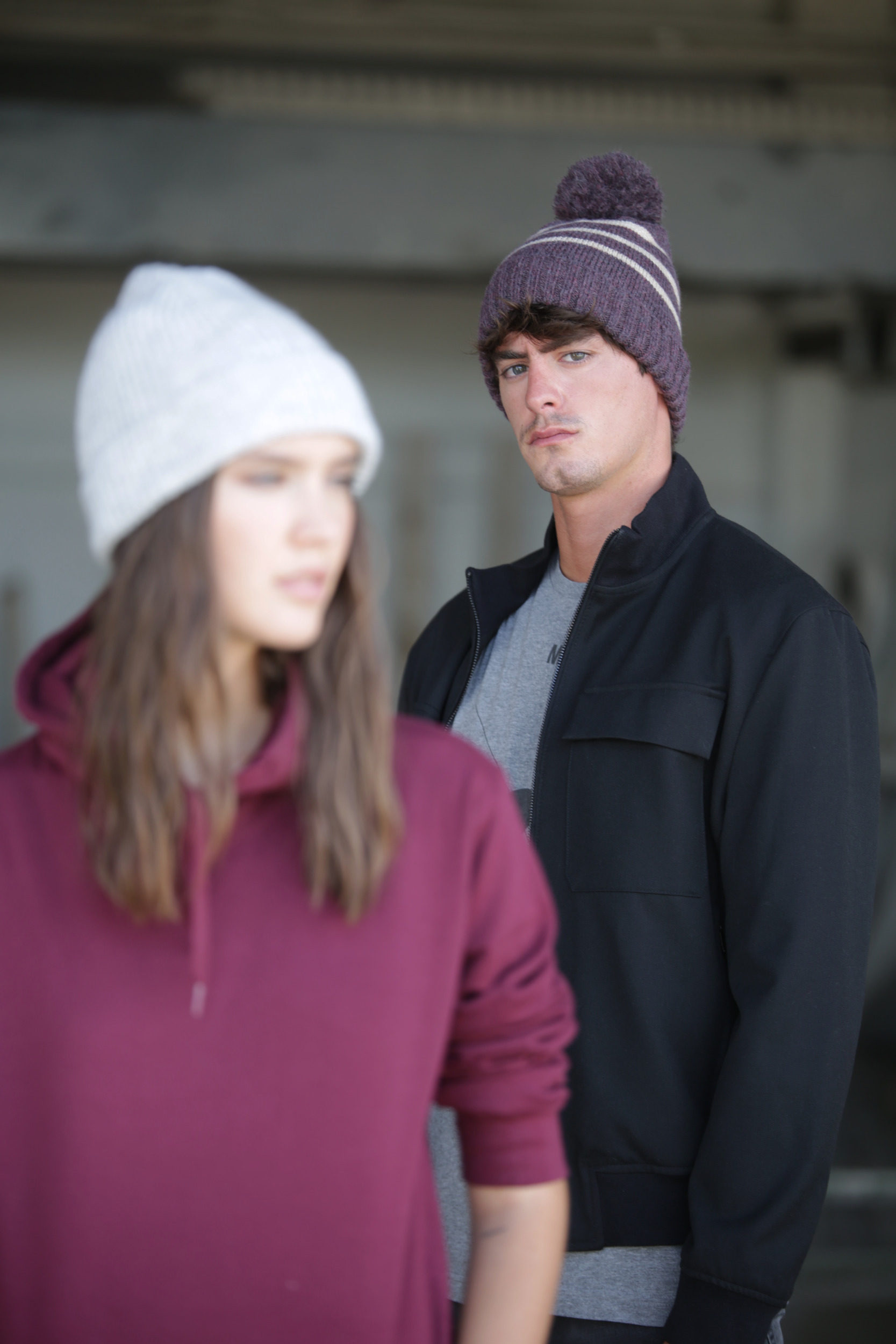 Hombre con gorro de lana y chaqueta negra mirando a cámara junto a mujer con sudadera y gorro desenfocada en primer plano