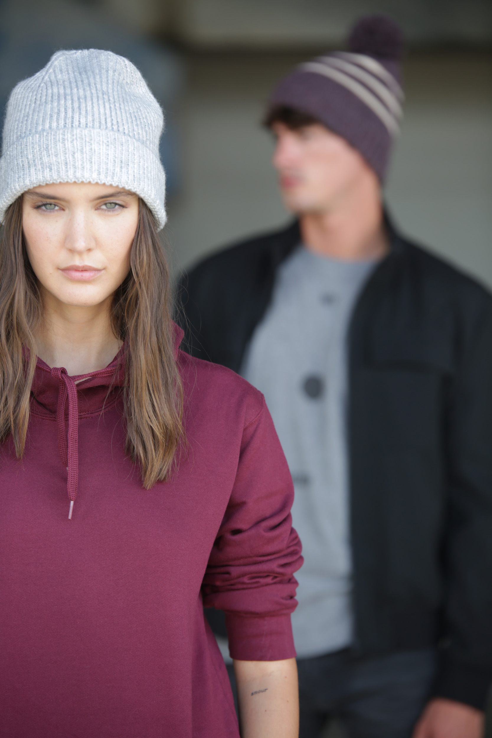 Primer plano de una mujer con gorro de punto gris y sudadera granate, con un hombre desenfocado detrás vestido con gorro y chaqueta.