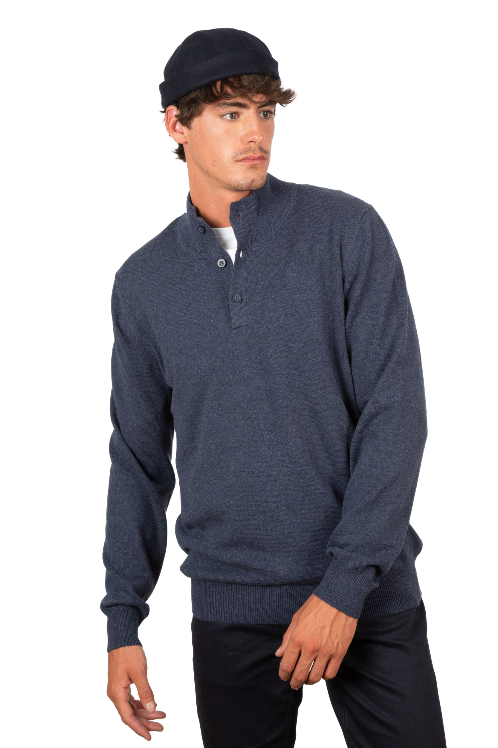 Hombre joven posando sobre fondo blanco con jersey azul marino de cuello alto y botones y gorro oscuro