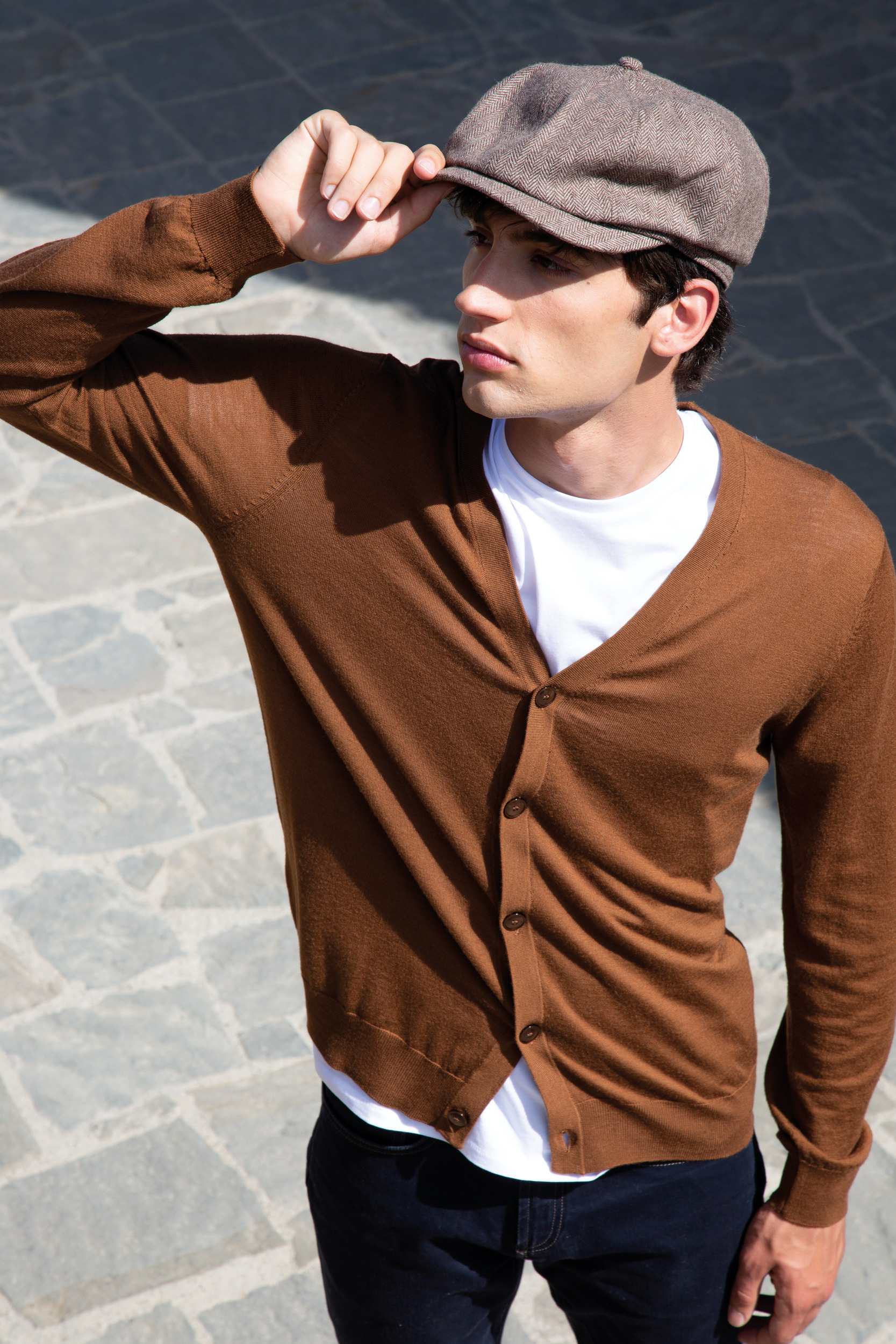 Hombre joven con cárdigan marrón y gorra plana en calle adoquinada mirada al horizonte