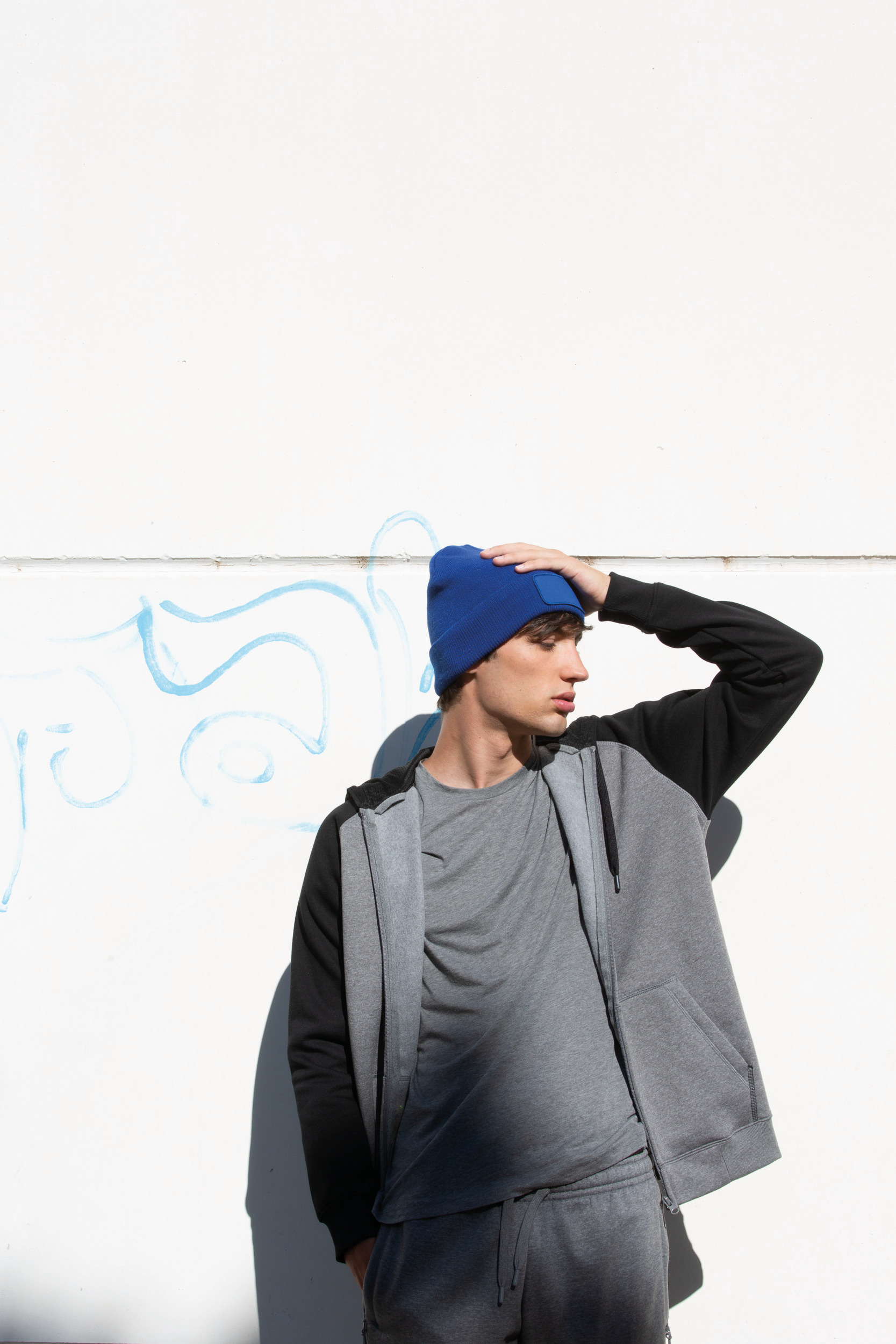 Joven con gorro de punto azul y chándal gris apoyado en una pared blanca con grafiti.