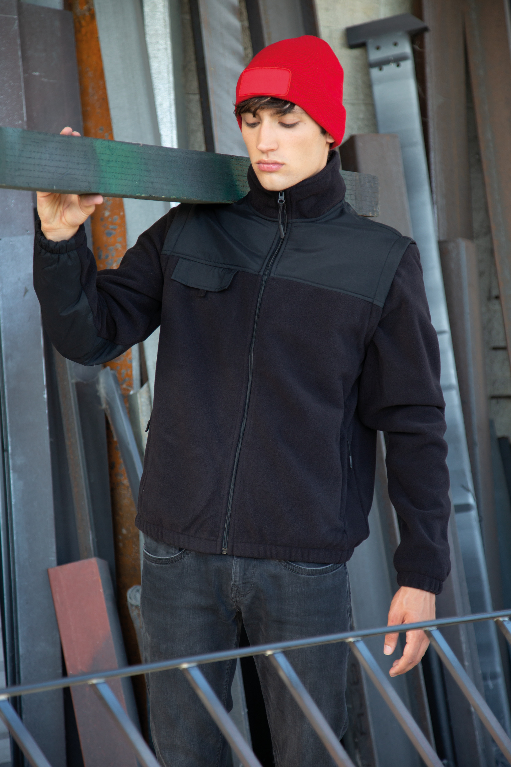 Hombre joven con gorro rojo y chaqueta negra apoyando una viga sobre el hombro en un taller metálico