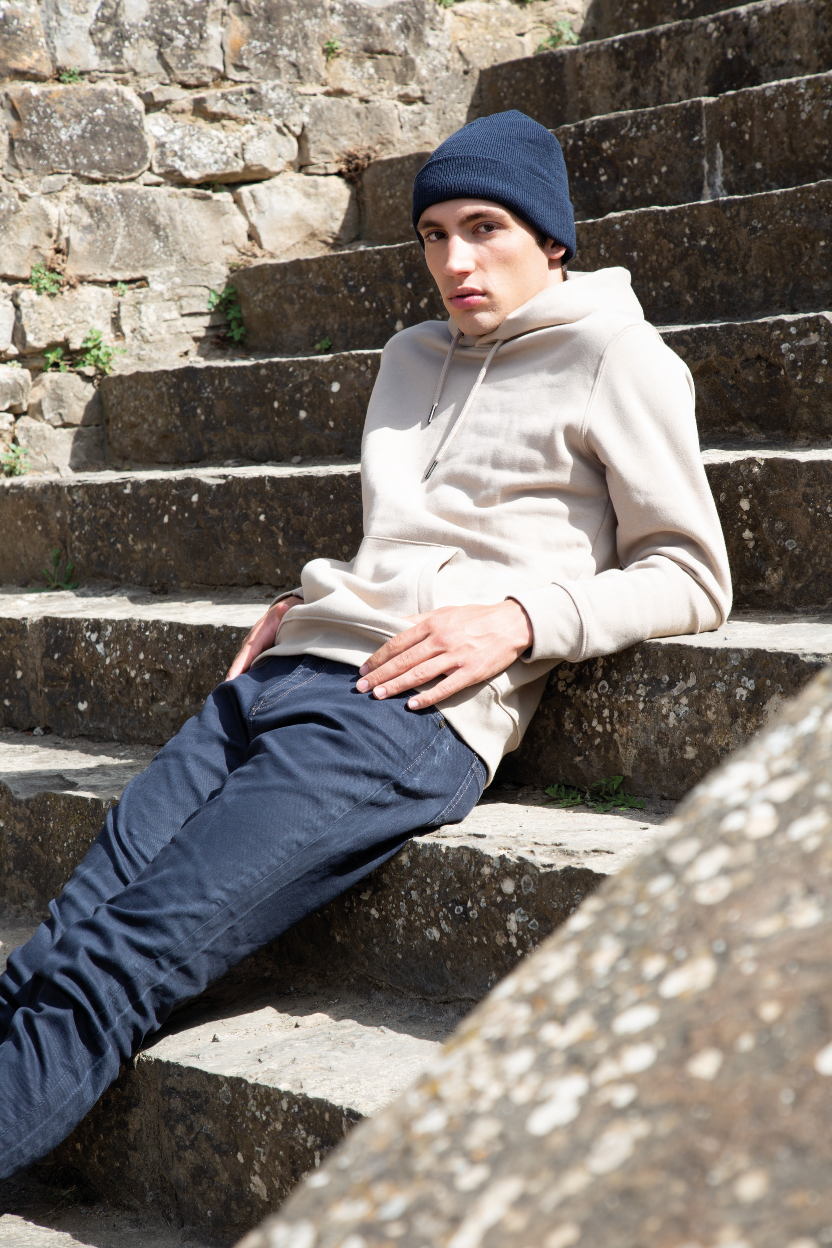 Joven con gorro azul y sudadera beige recostado en escalones de piedra al aire libre
