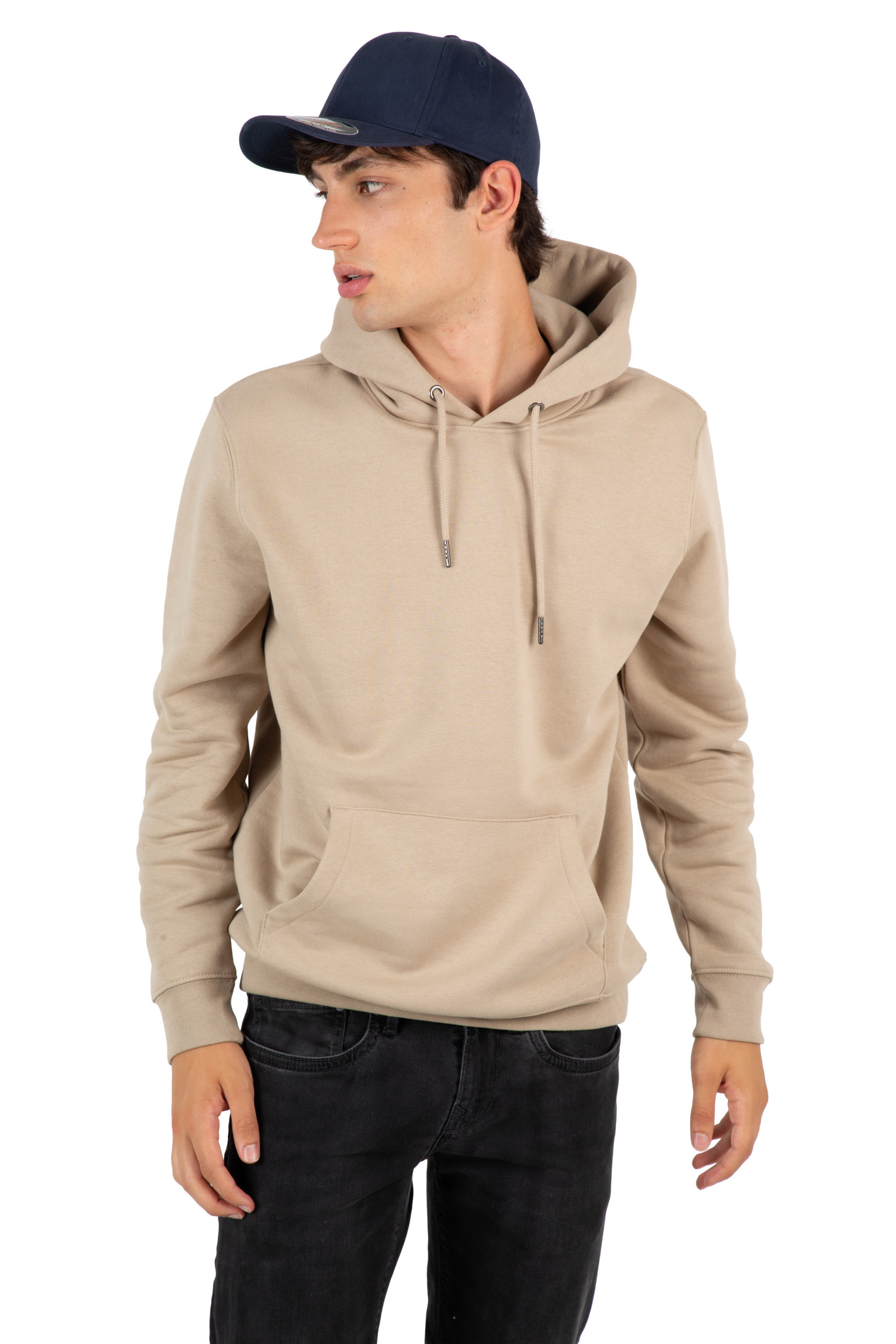 Joven de perfil con gorra azul marino, sudadera con capucha beige y vaqueros negros sobre fondo blanco.