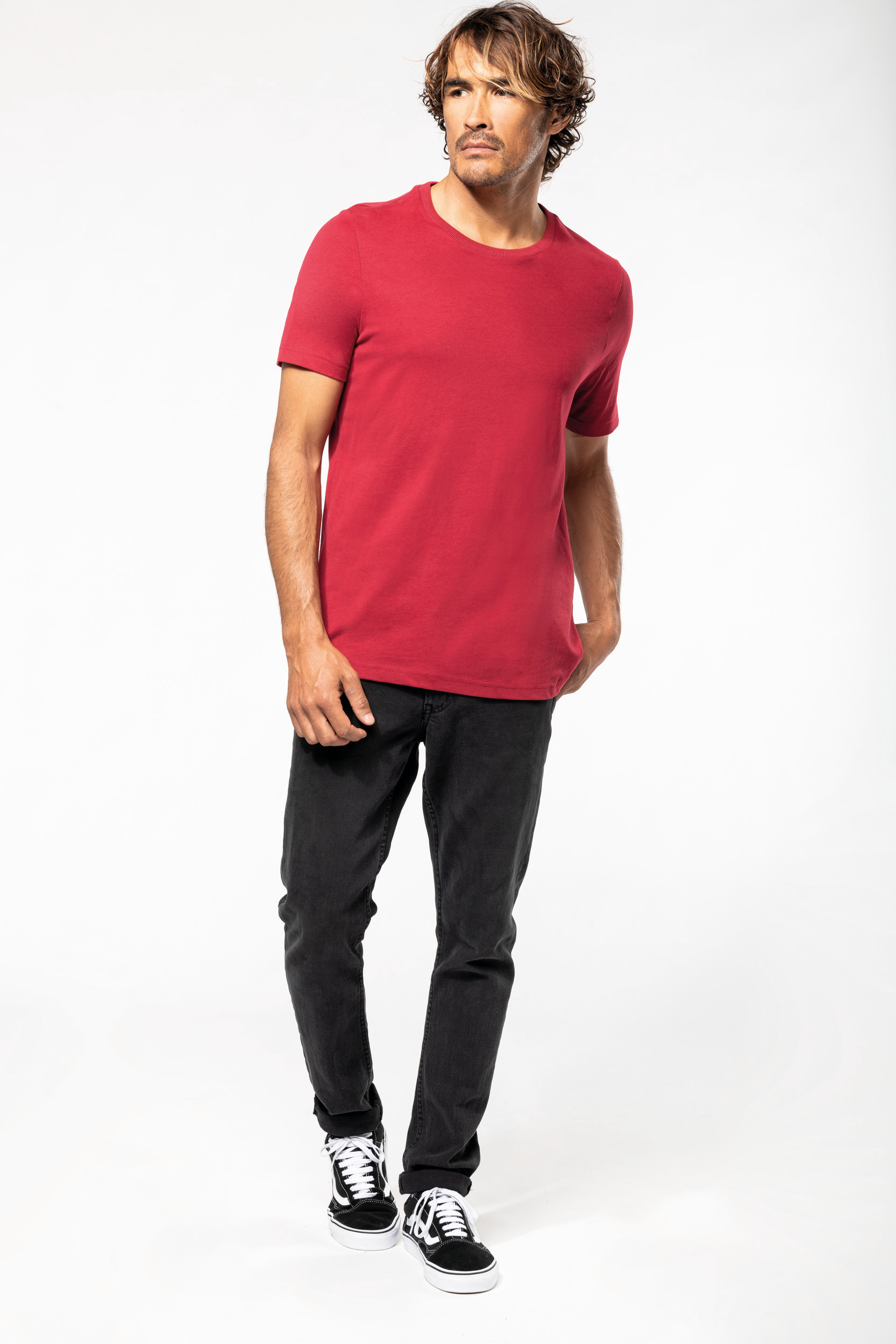 Hombre de pie con camiseta roja, vaqueros negros y zapatillas sobre fondo blanco.