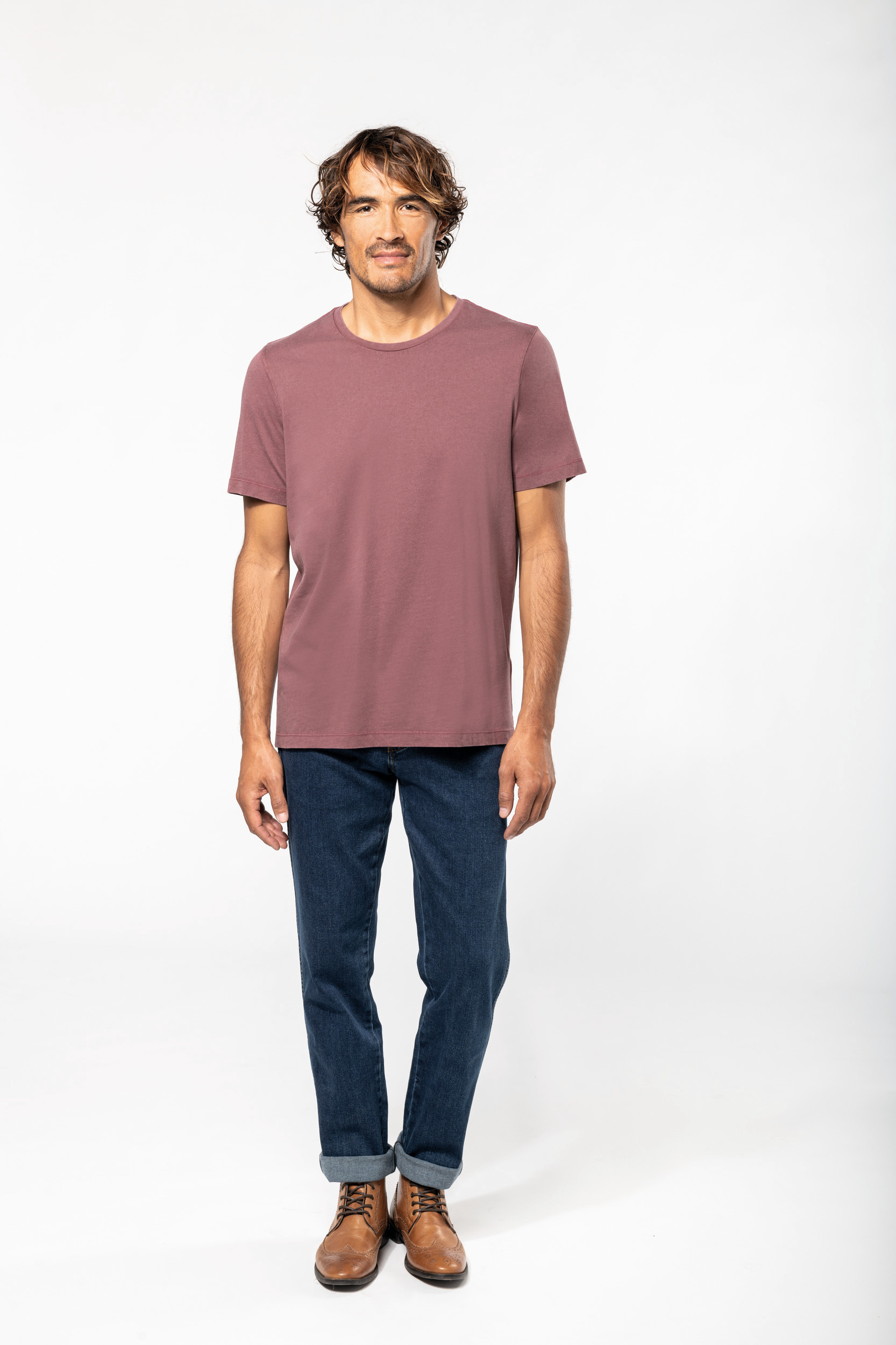Hombre joven de pie con camiseta rosa y vaqueros oscuros sobre fondo blanco.