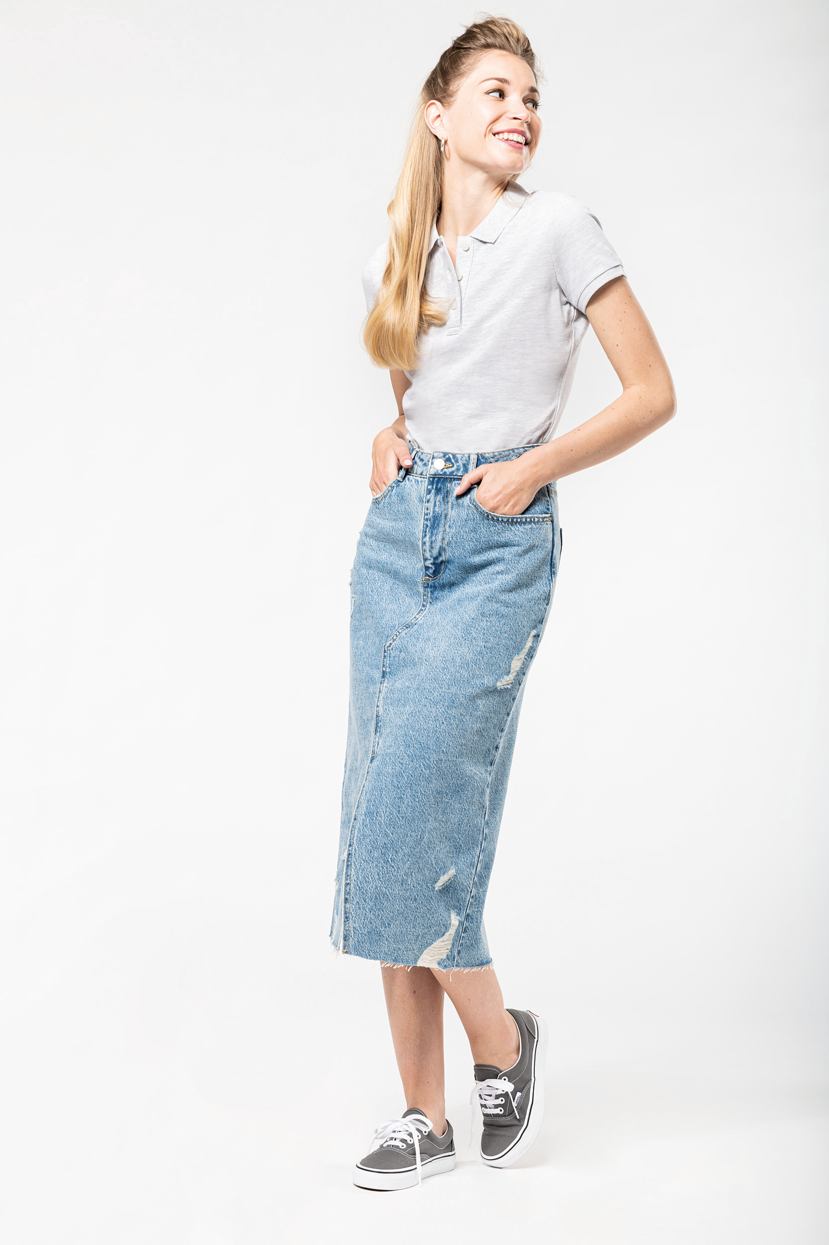 Mujer joven sonriente con polo blanco, falda vaquera larga y zapatillas grises sobre fondo blanco.