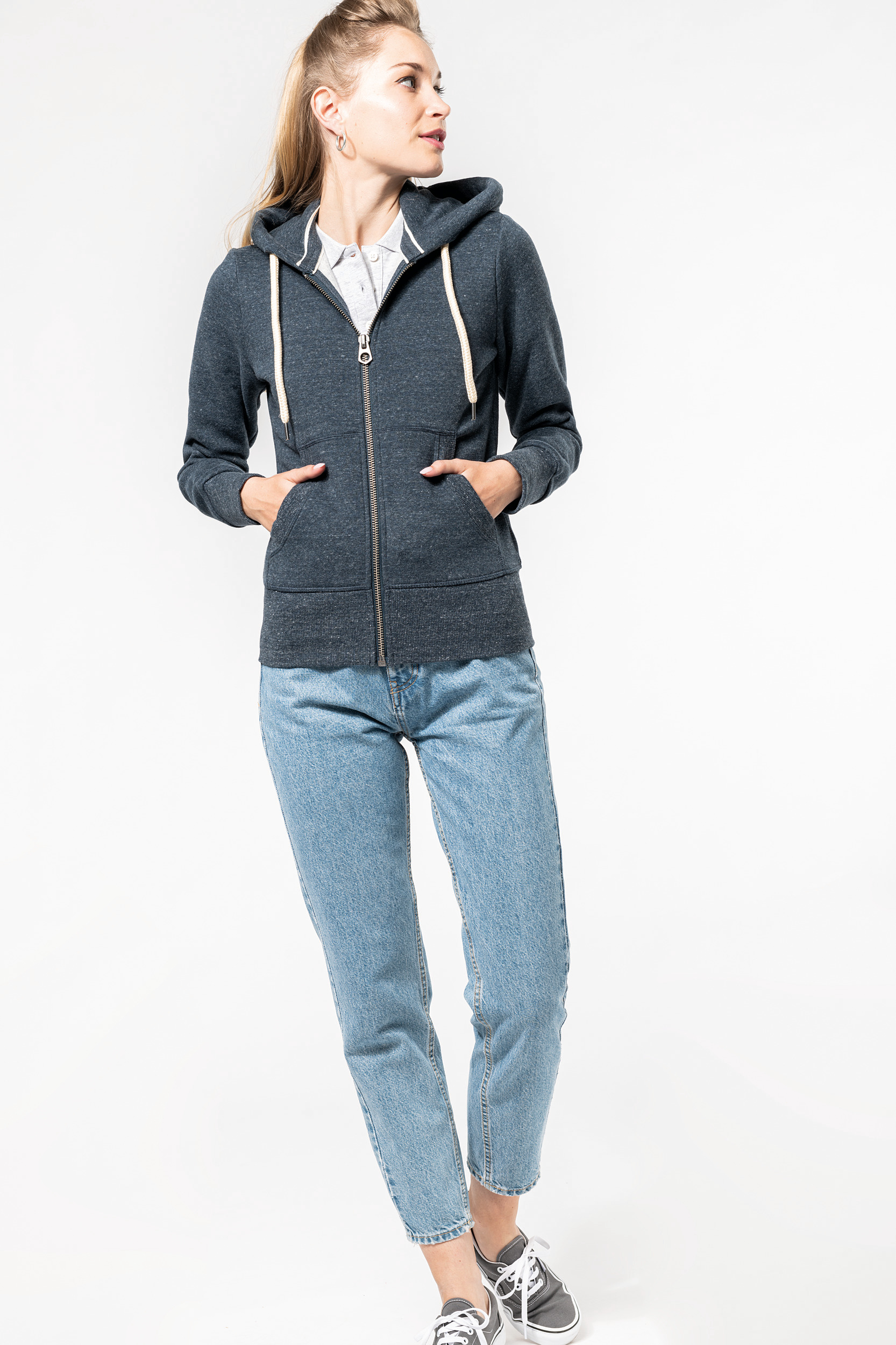 Mujer joven con sudadera con capucha azul marino, vaqueros y zapatillas grises sobre fondo blanco.