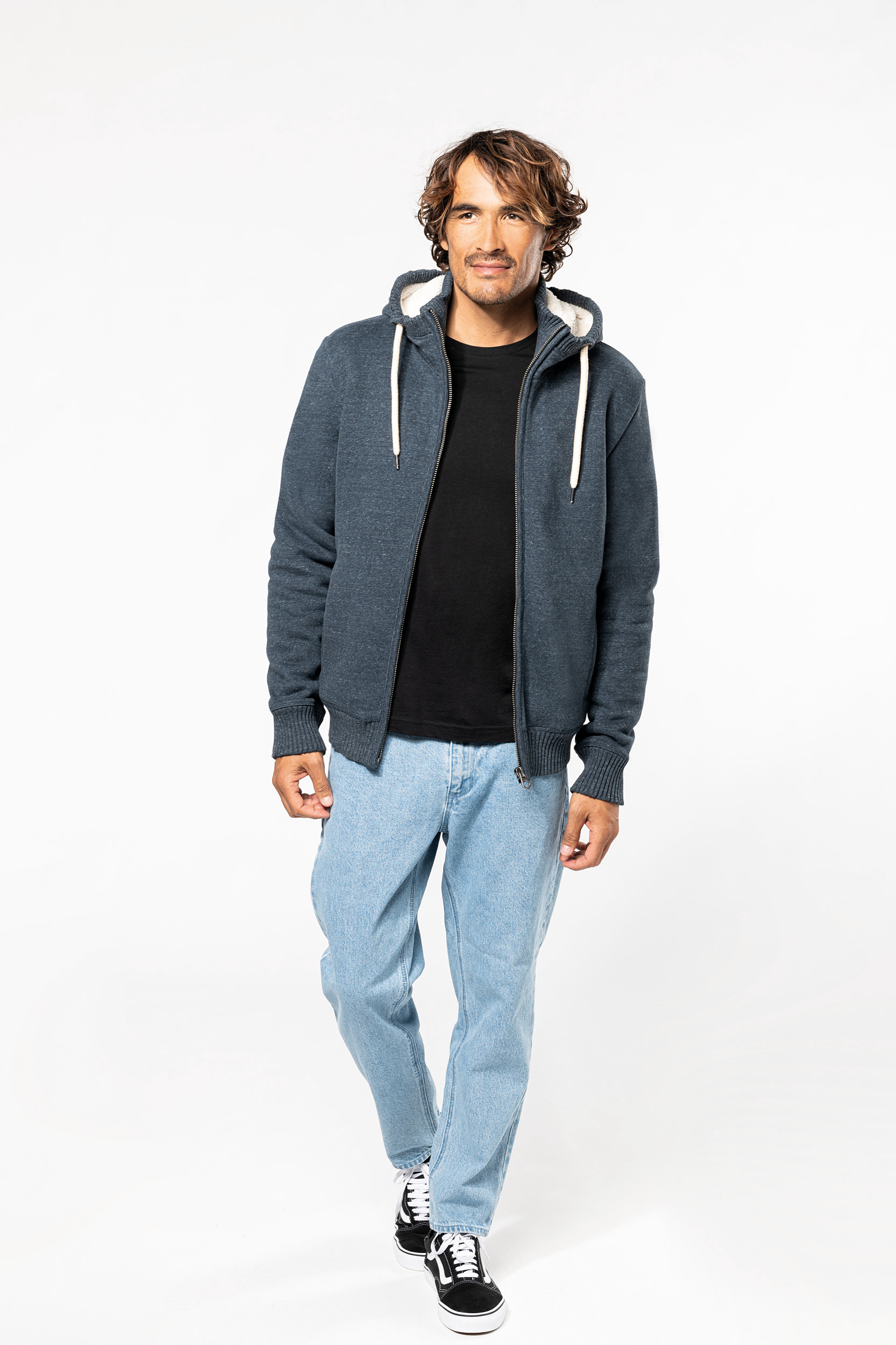 Hombre posa con sudadera azul con capucha, camiseta negra y vaqueros claros sobre fondo blanco